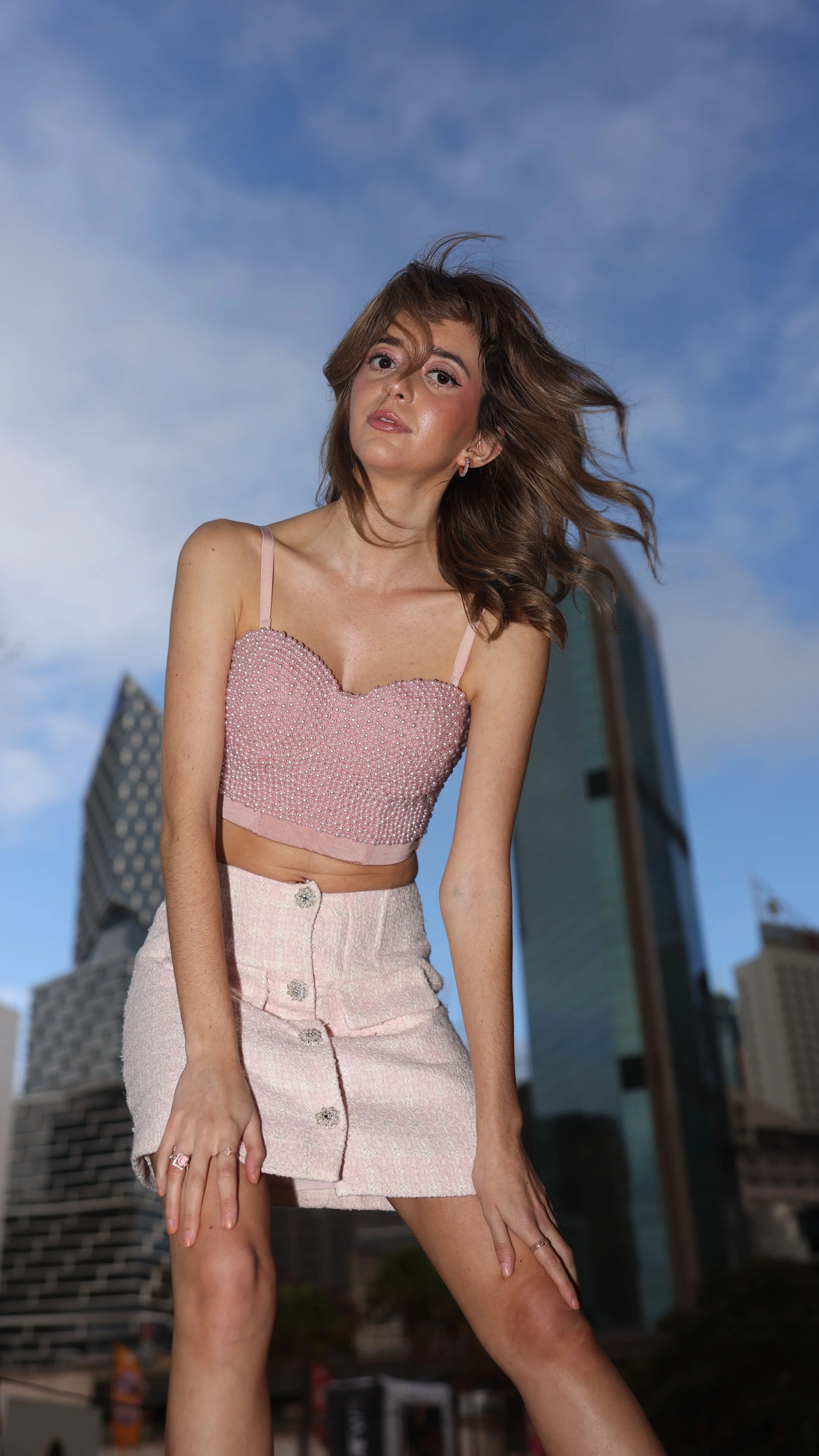 A woman with wavy brown hair wearing a pink embellished crop top and matching high-waisted skirt posing outdoors in front of city buildings with a blue sky.