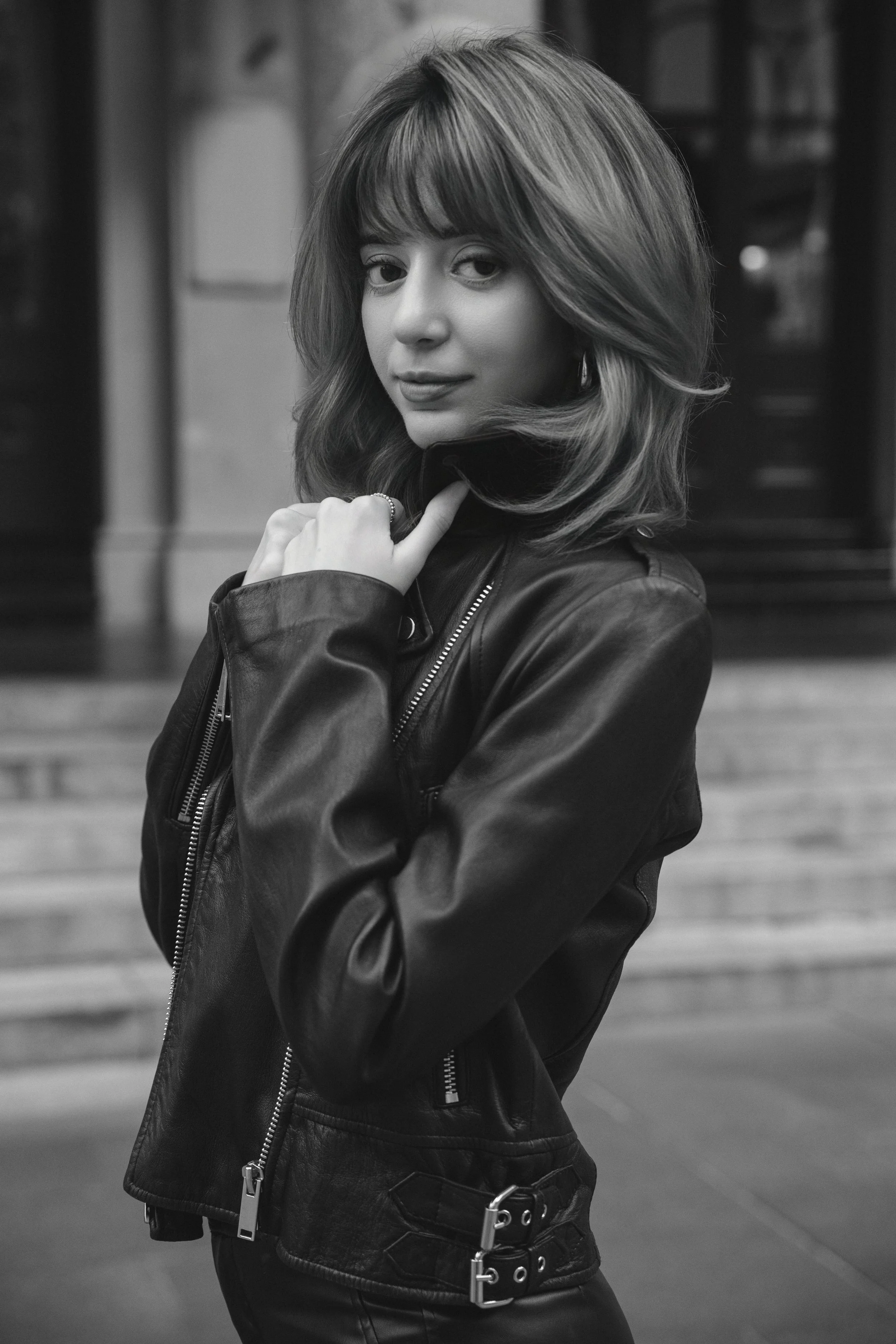 A woman with shoulder-length hair, wearing a black leather jacket, stands in an outdoor urban setting. She has her hand near her face and looks at the camera.