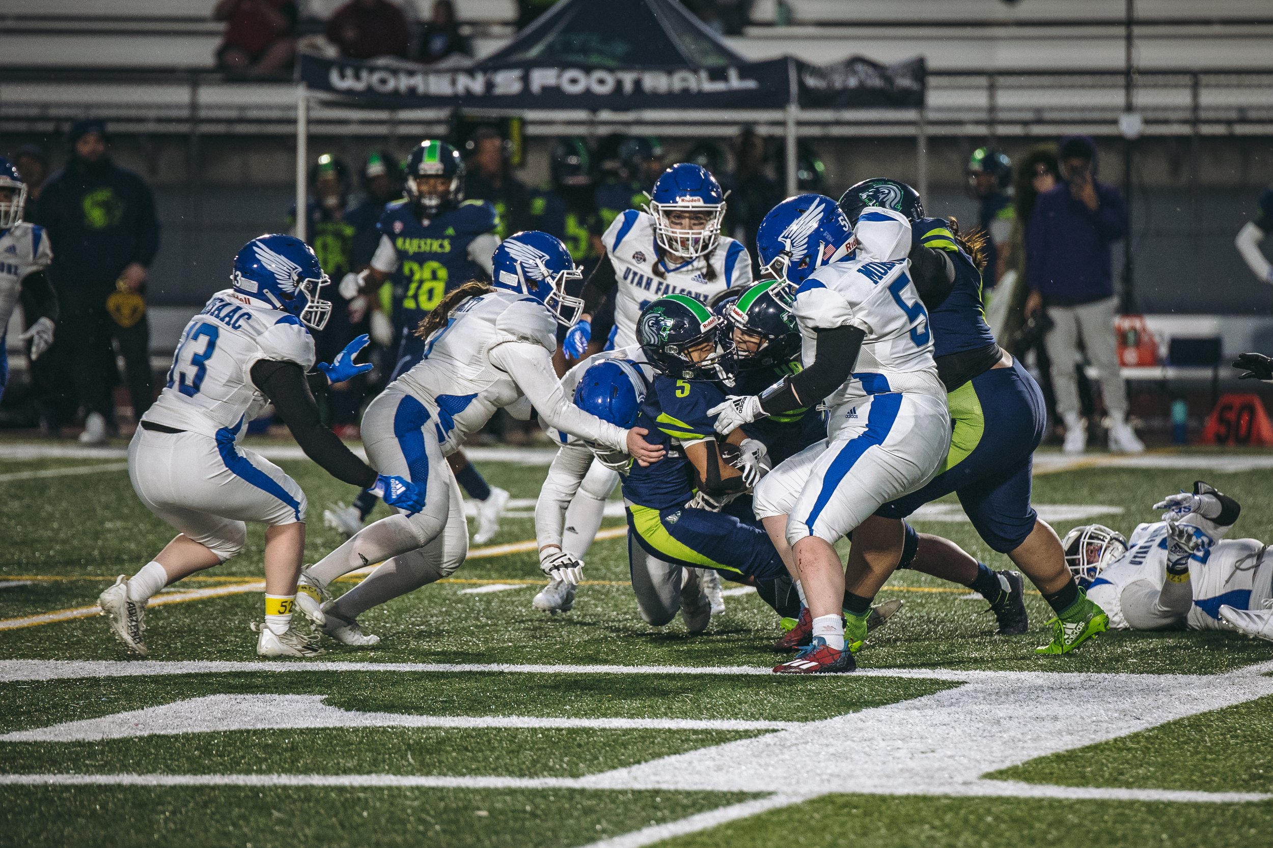A group of football players during a game, with players from the white and blue team tackling the player from the navy and green team on the field.