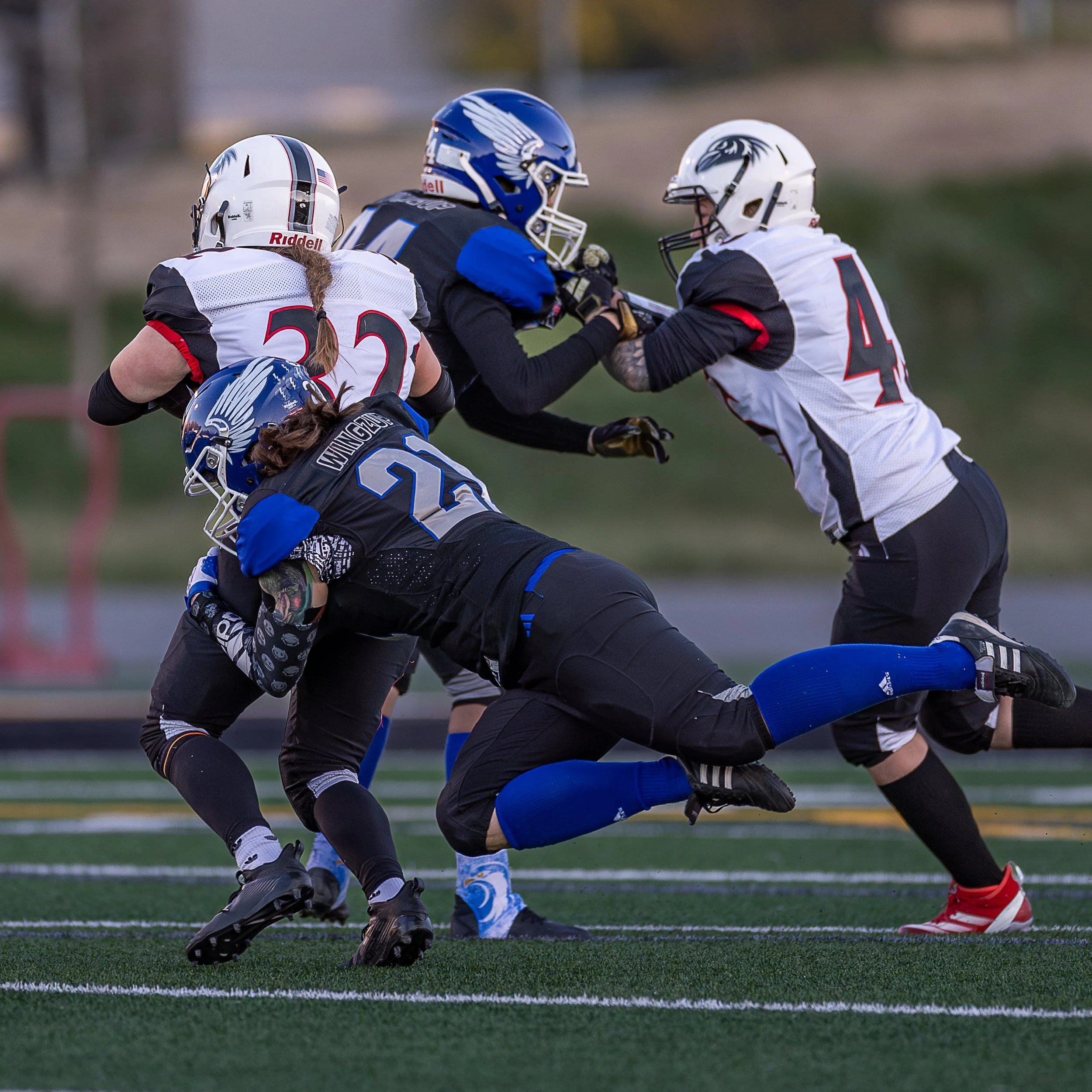 A group of football players in black, white, and blue uniforms engaging in a play on a field, with one player tackling another.