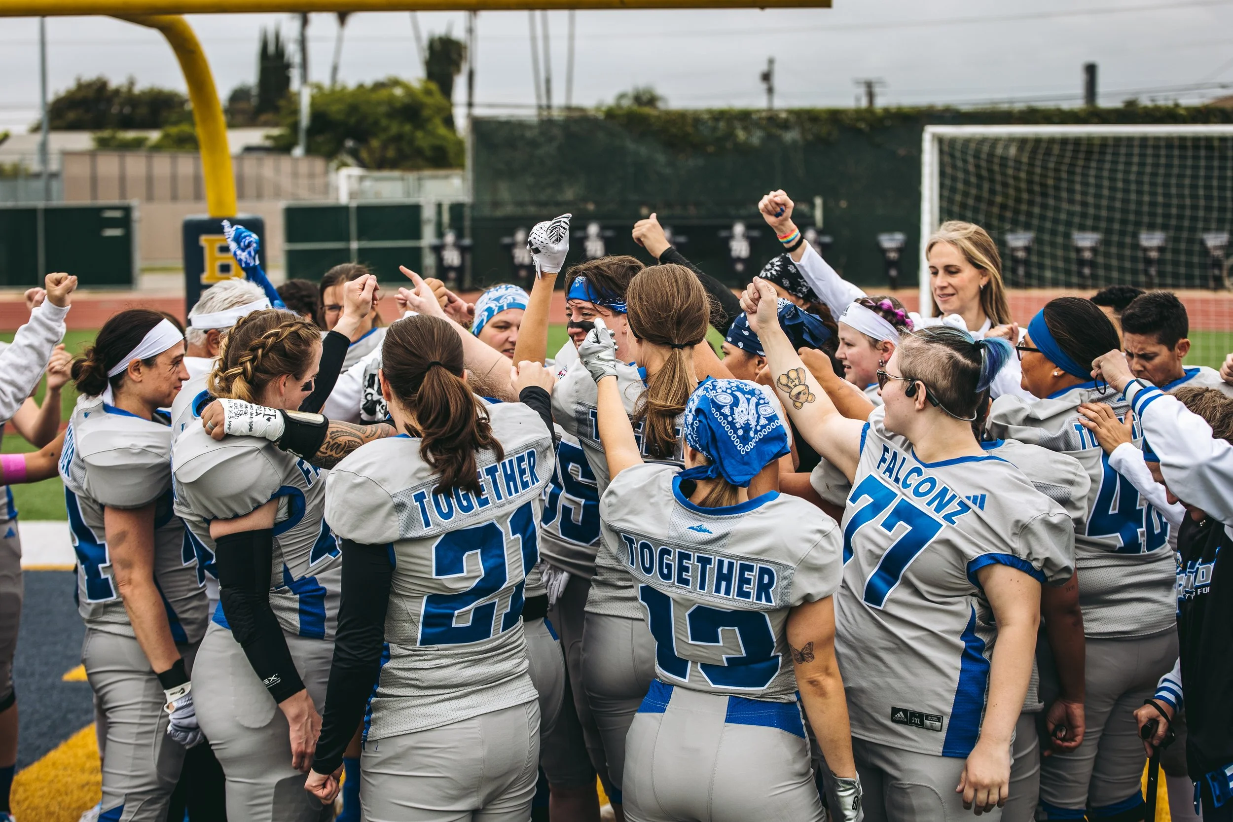 A group of female football players wearing gray jerseys with blue accents and headbands, celebrating and cheering together on a football field during daytime.