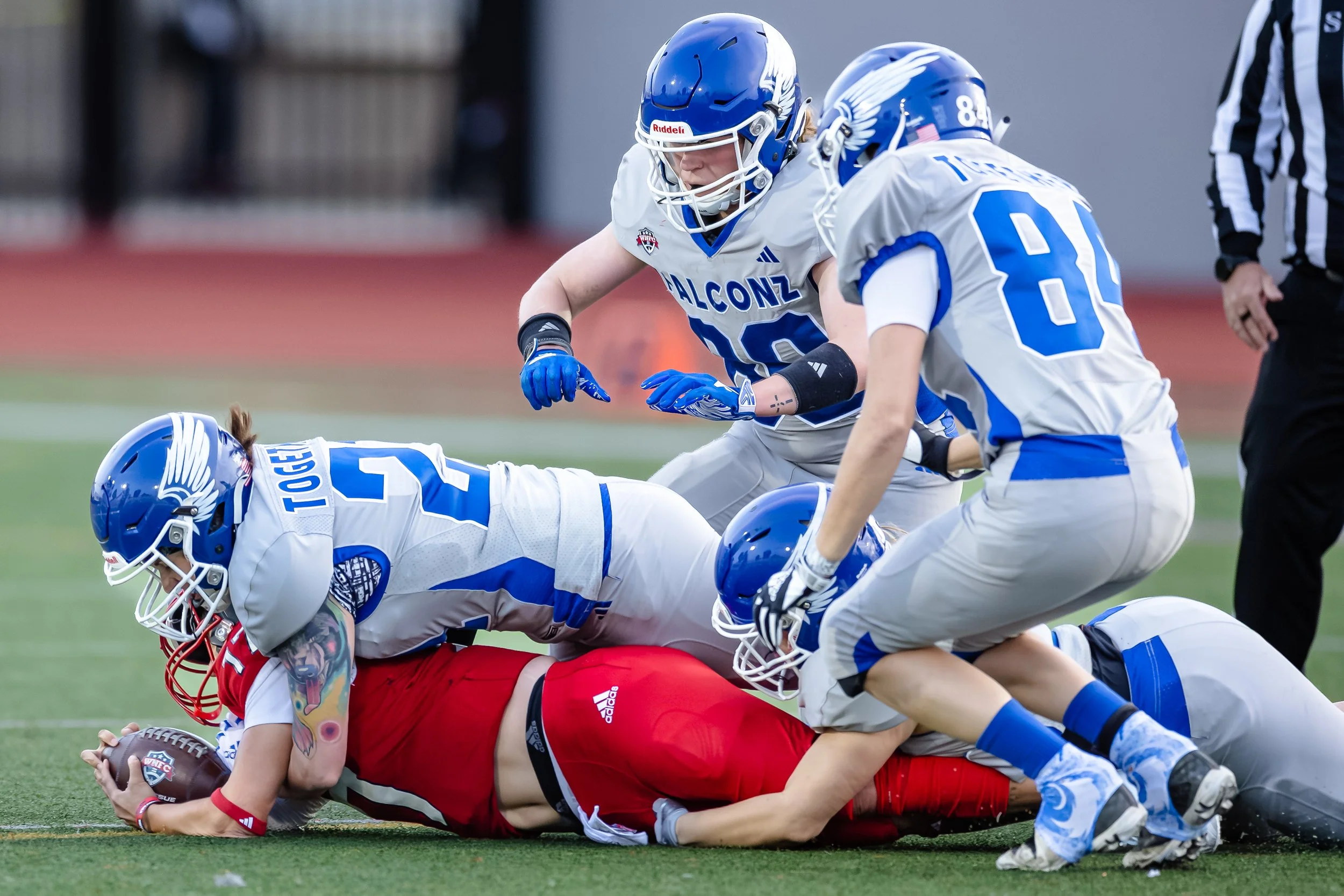 American football players in action, with a player in red holding the football on the ground while being tackled by players in white and blue uniforms on a green field.
