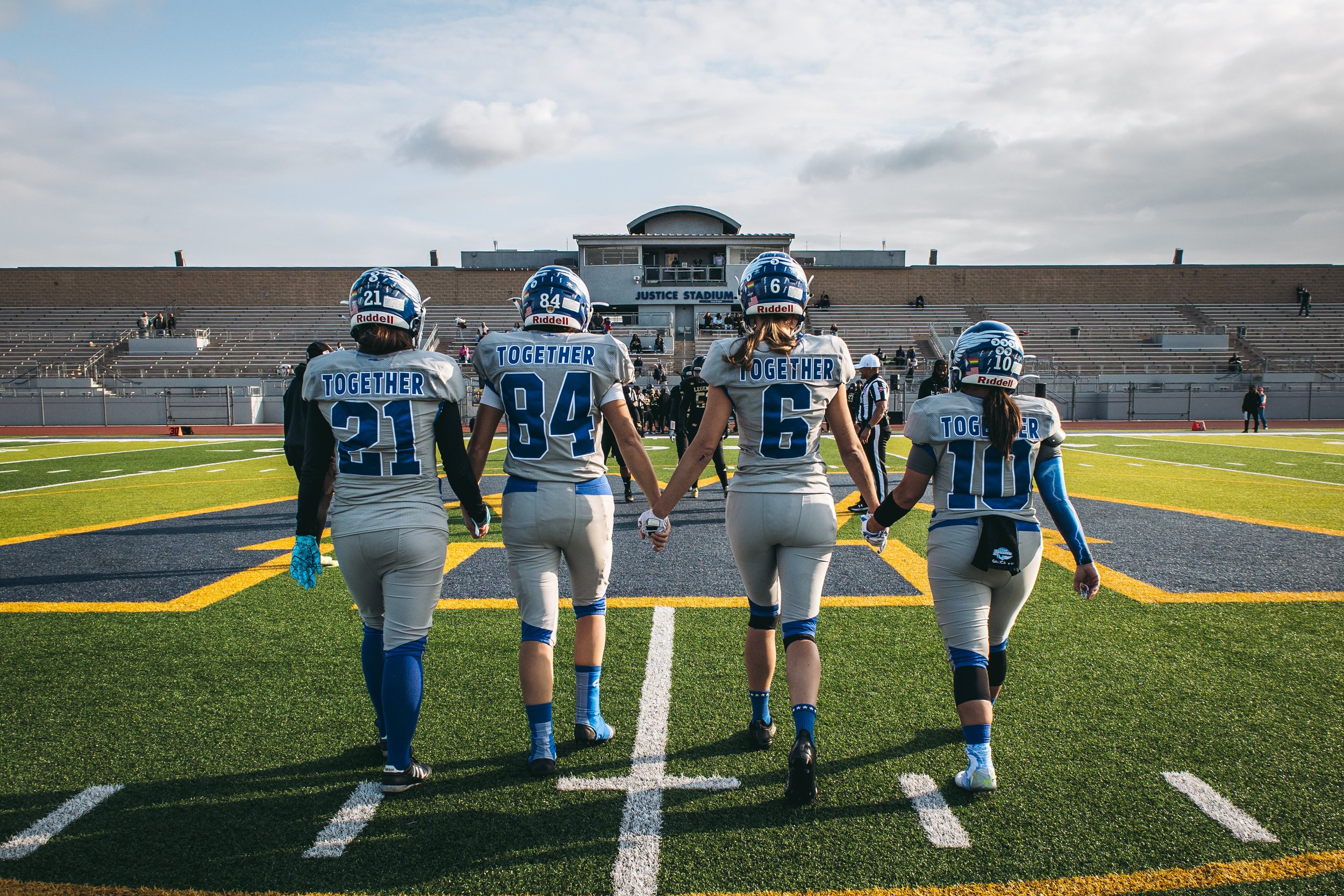 Group of female American football players walking onto the field holding hands, wearing uniforms with the word 'Together' on the back, at a stadium with empty stands and a cloudy sky.