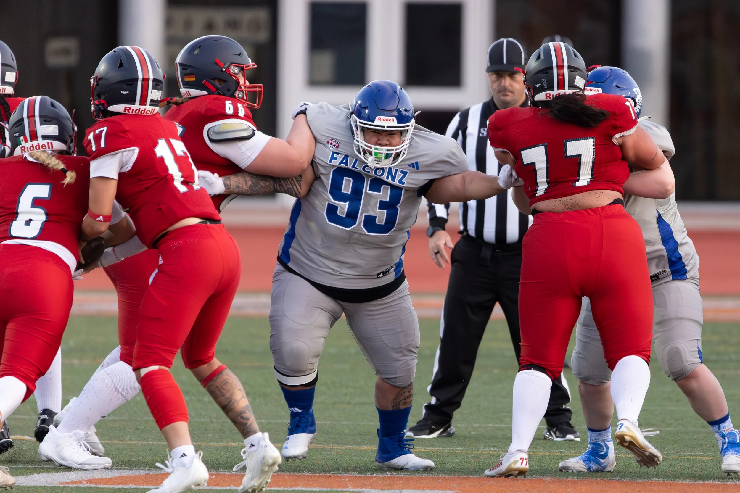American football game with players in red and gray uniforms engaging at the line of scrimmage, an official in black and white striped uniform standing behind them.