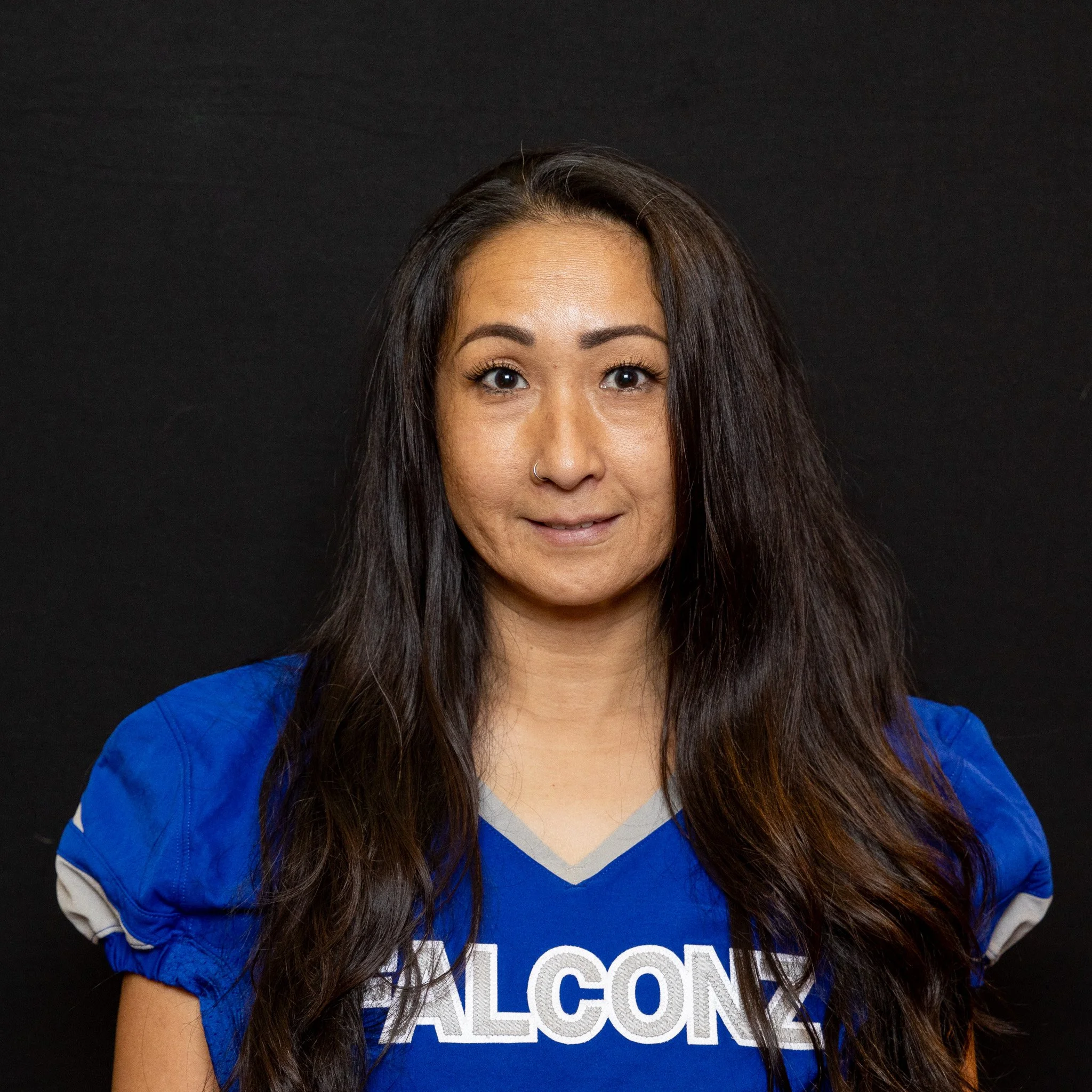 A woman with long dark hair and a nose piercing wearing a blue sports jersey with white and gray accents, standing against a black background.