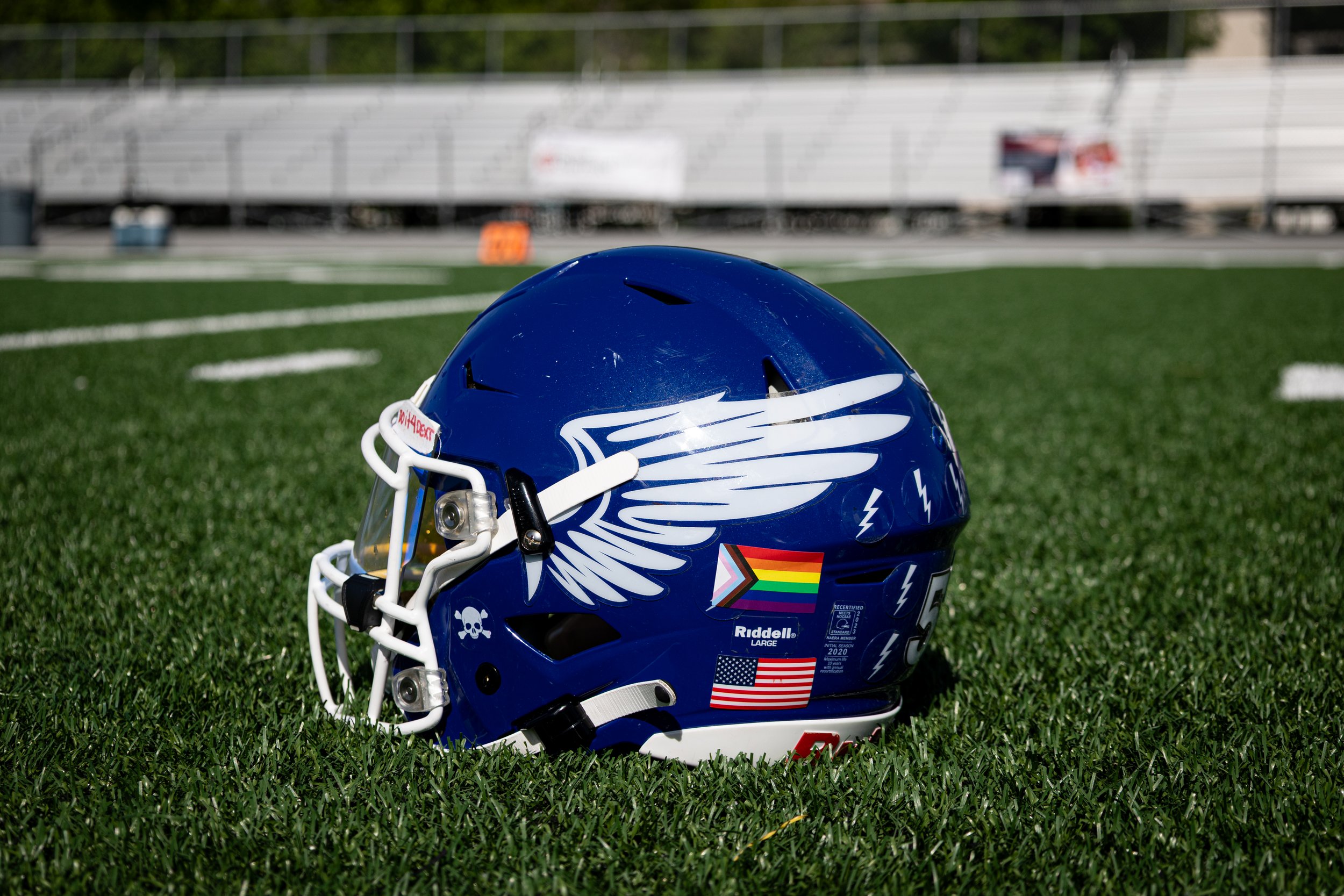 A blue football helmet with white and black decals, including an angel wing design, rainbow pride flag, American flag, and a skull sticker, placed on a green football field.