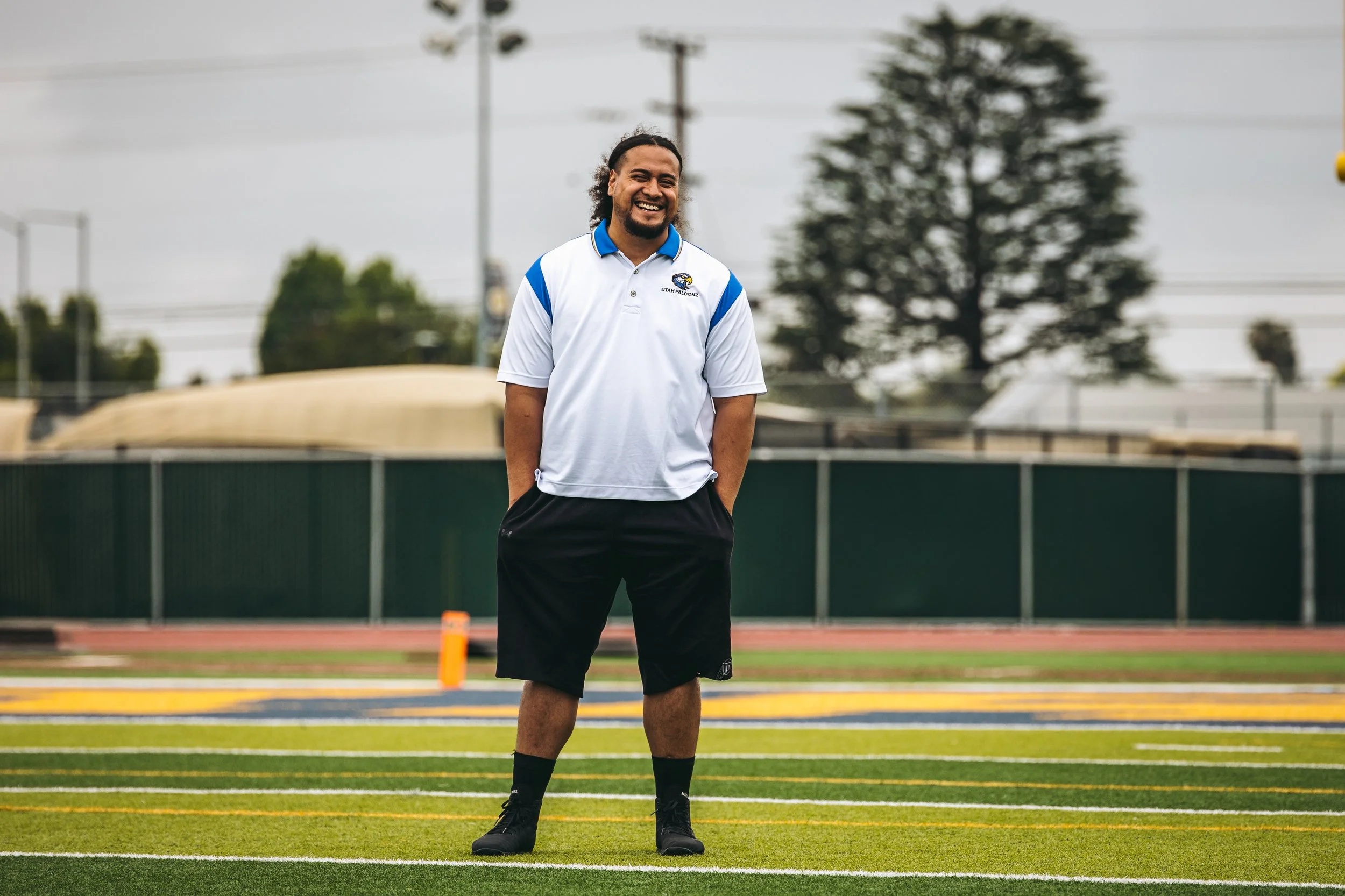 A man in a white and blue shirt and black shorts standing on a football field, smiling with hands in pockets.
