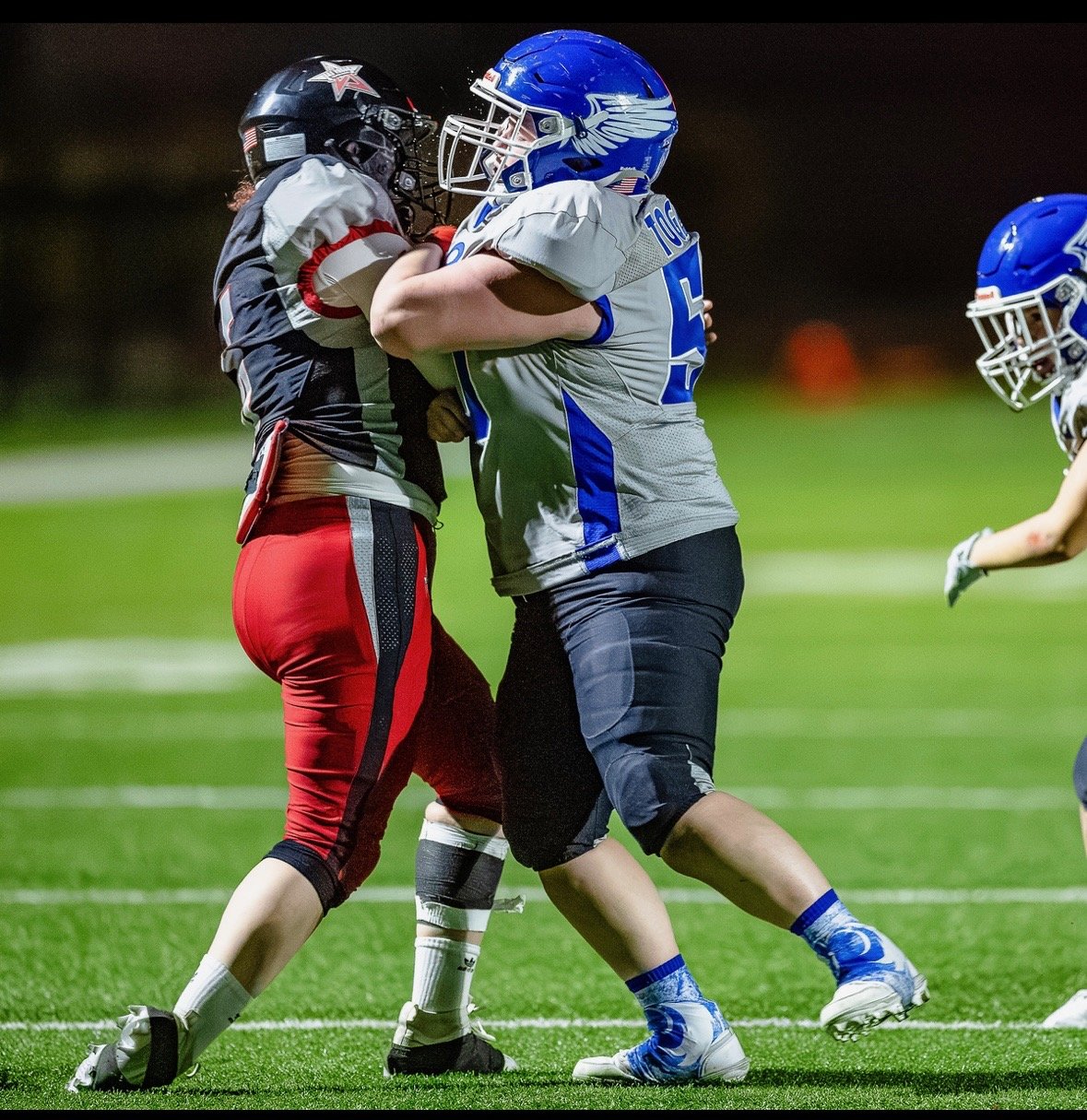 Two football players, one in a gray and blue uniform and the other in a black and red uniform, engaging in a tackle on the field at night.