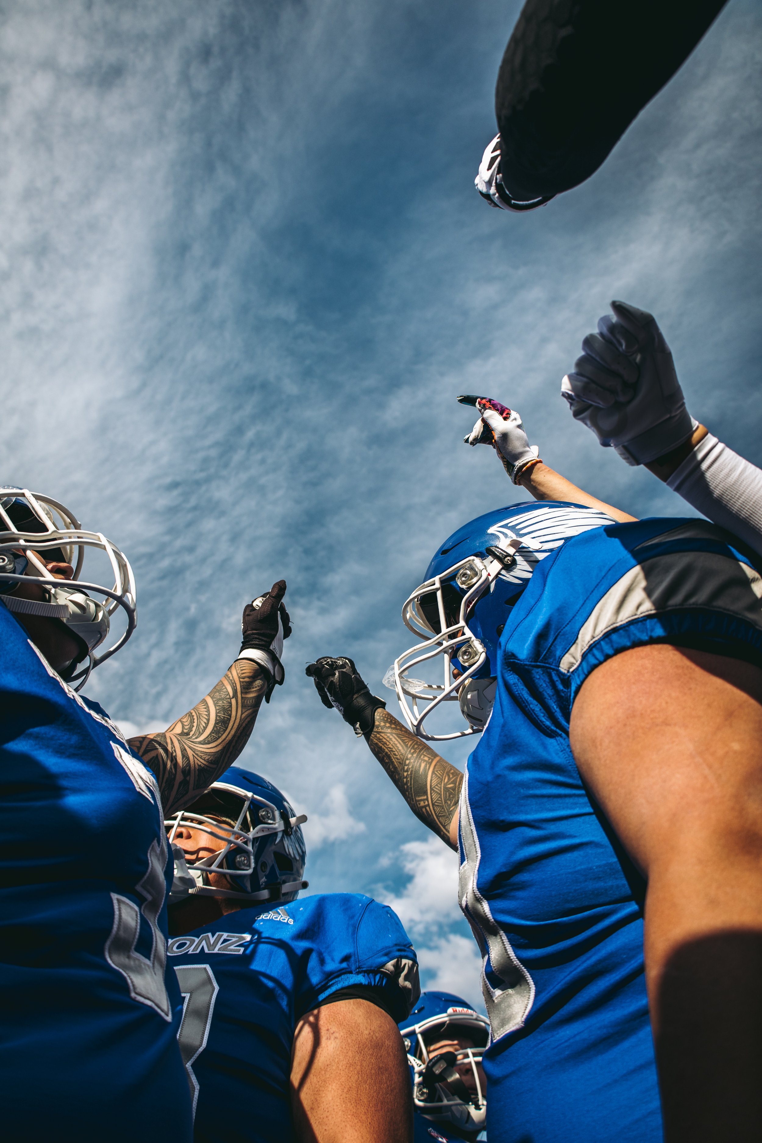A team of American football players wearing blue jerseys and helmets are gathered in a huddle, pointing upward with a clear blue sky overhead.