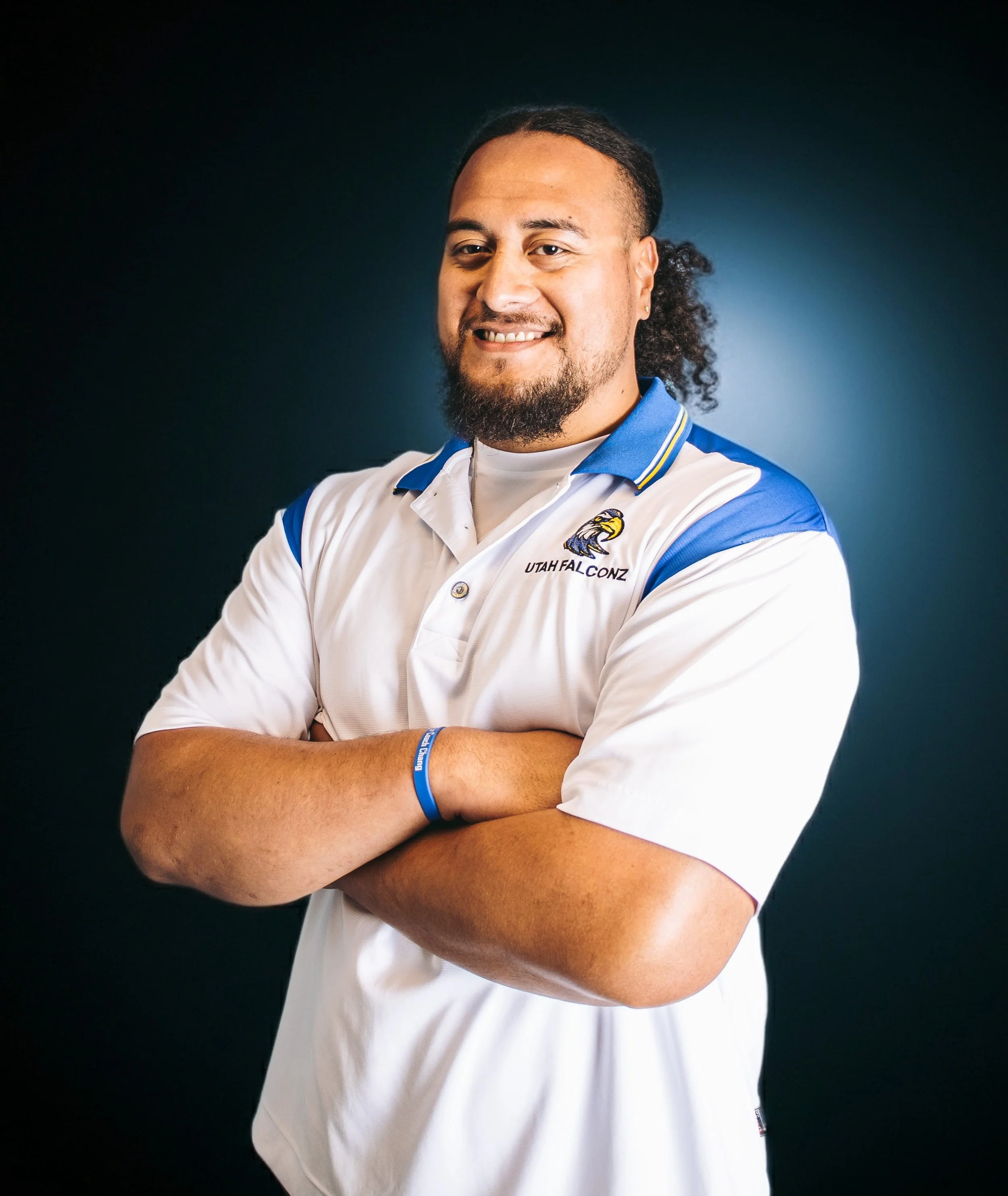 A man in a white and blue shirt and black shorts standing on a football field, smiling with hands in pockets.