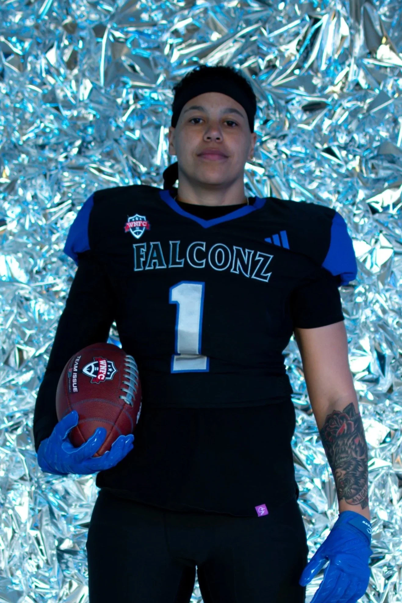 Young football player with shoulder-length hair wearing glasses, a blue football jersey with white text 'FALCONZ', and braces, smiling at the camera against a black background.