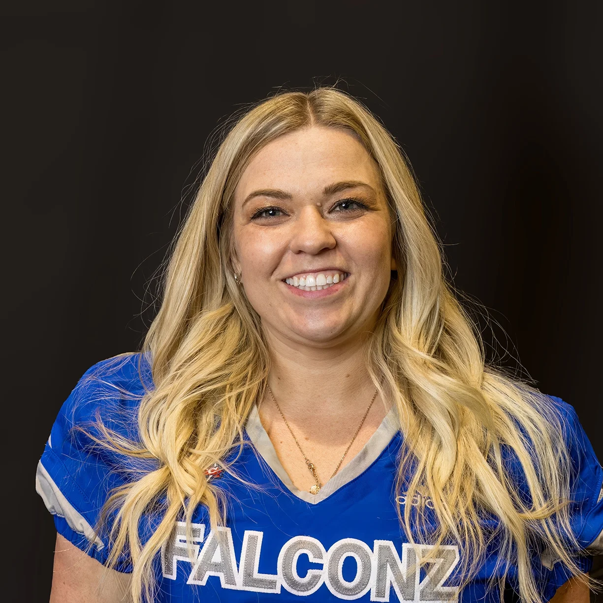 A woman with long blonde hair smiling, wearing a blue sports jersey with 'FALCONZ' written on it, against a plain dark background.