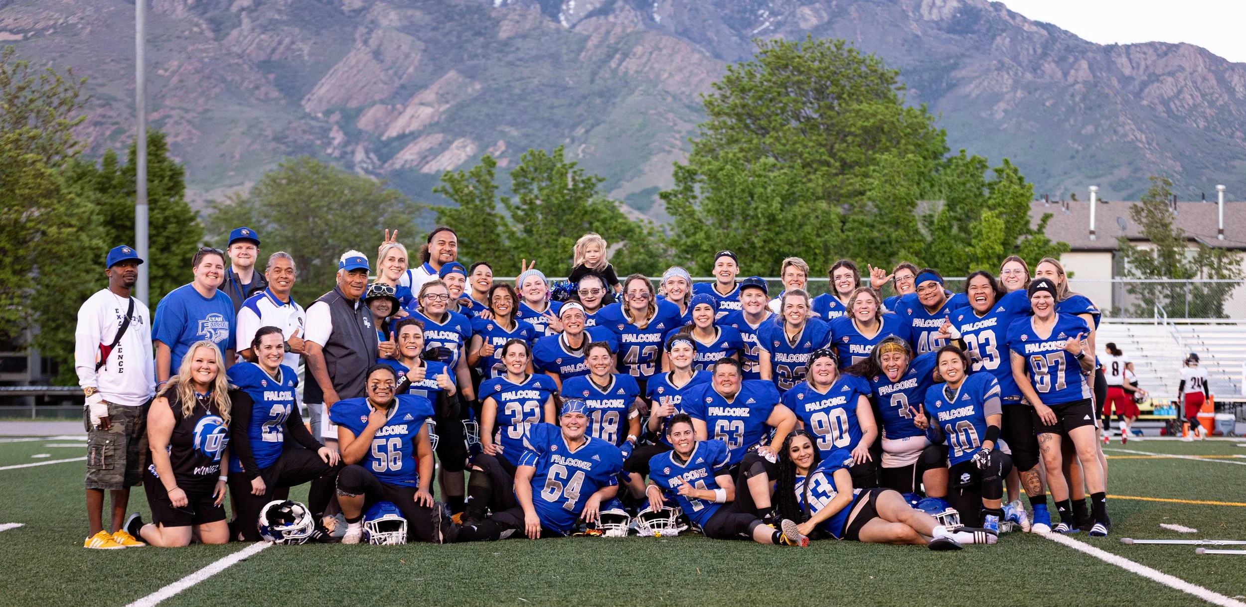 Group of female and male football players and coaches on a football field, wearing blue and black uniforms, smiling and posing for a team photo.