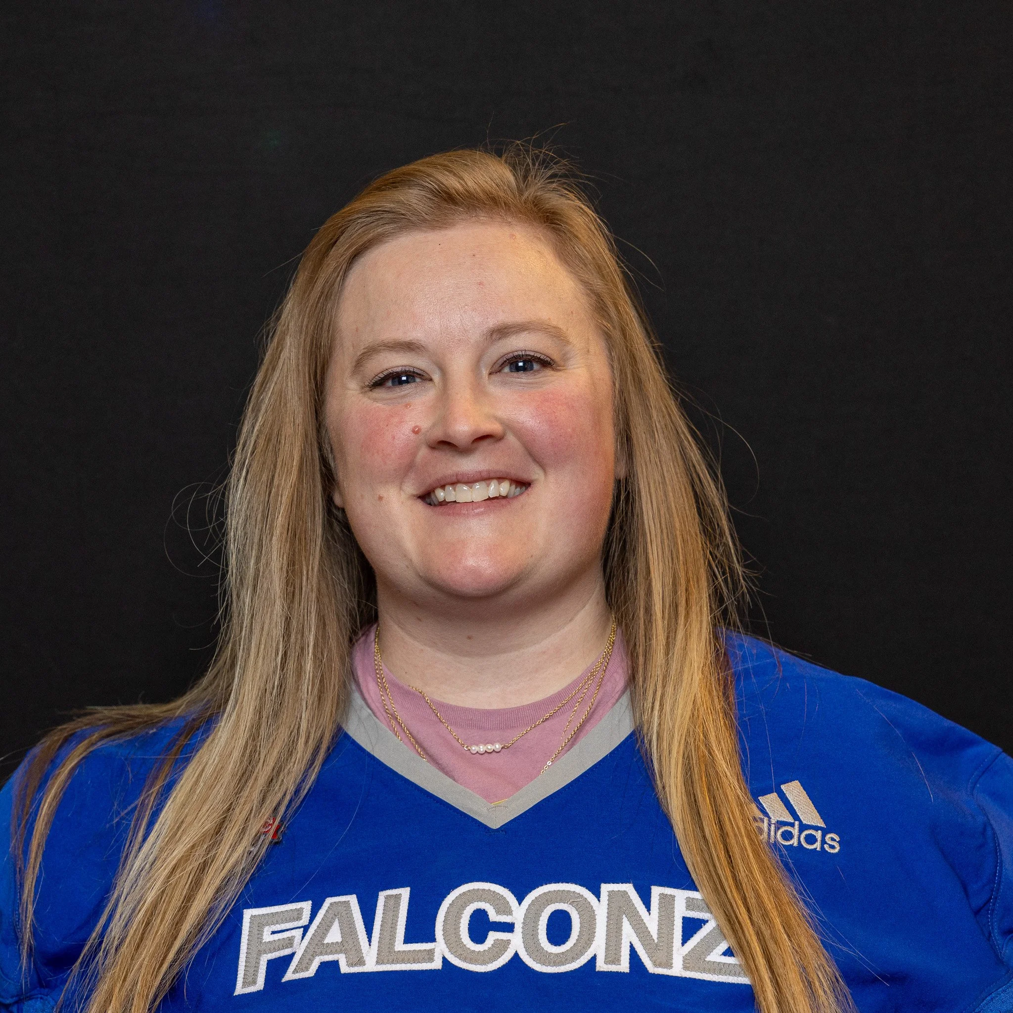 A woman with long red hair wearing a blue sports jersey with 'FALCONZ' on it and smiling against a black background.