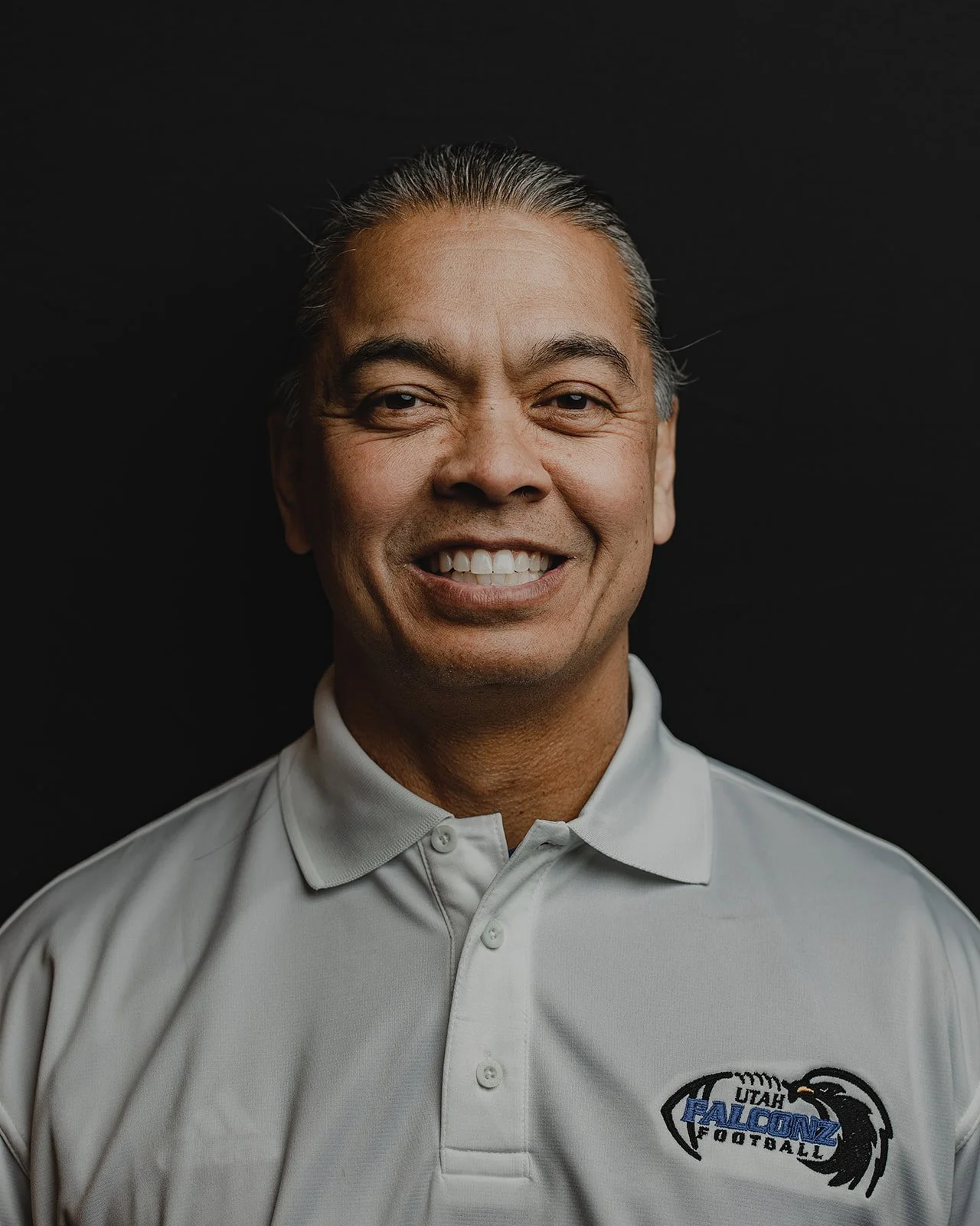 A smiling man with gray hair wearing a white polo shirt with a logo that reads 'Utah Falcons Football' on a black background.