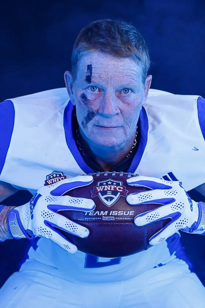 A woman with long red hair wearing a blue sports jersey with 'FALCONZ' on it and smiling against a black background.