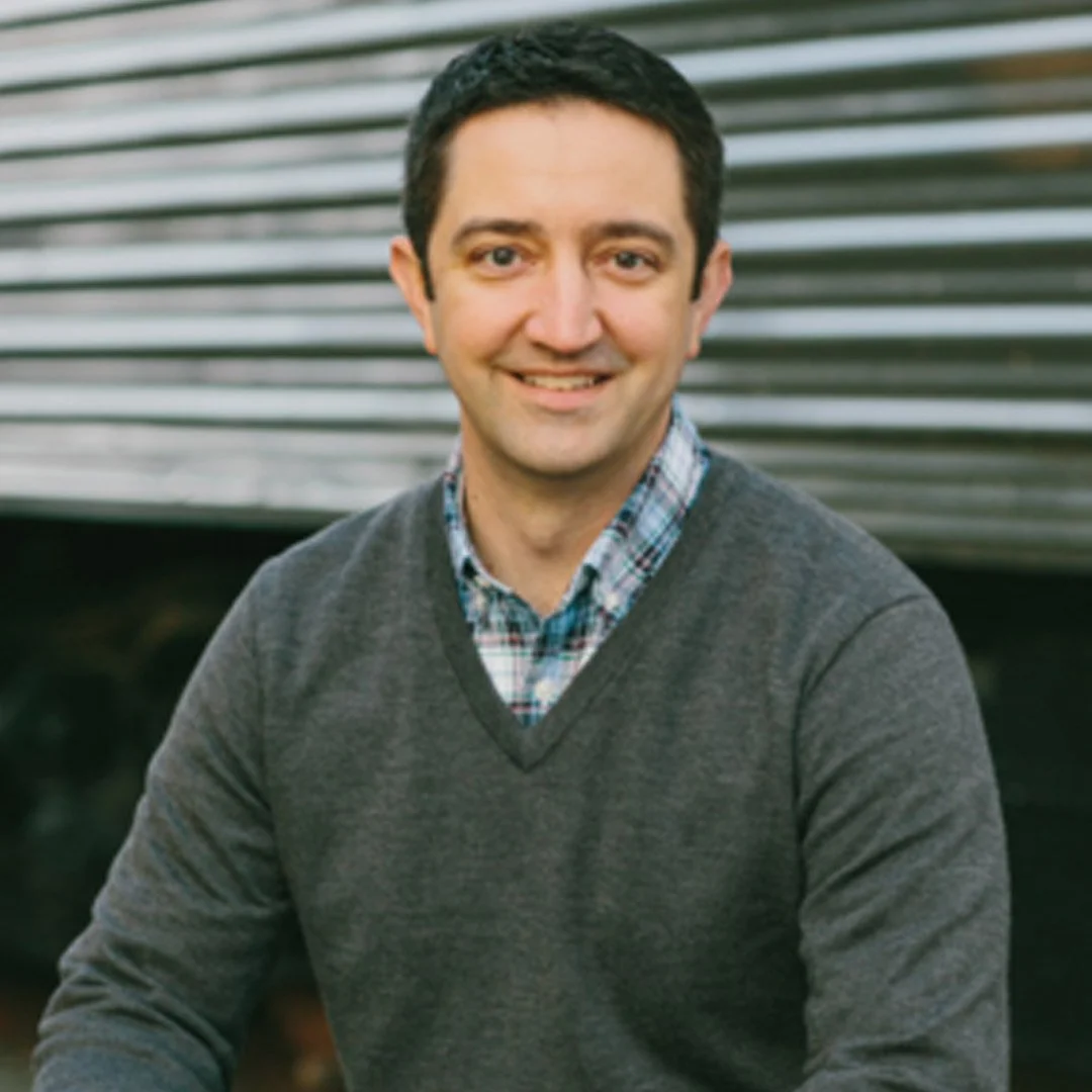 A man with short black hair and light skin, smiling, wearing a plaid shirt under a gray sweater, sitting outdoors in front of a metal building.