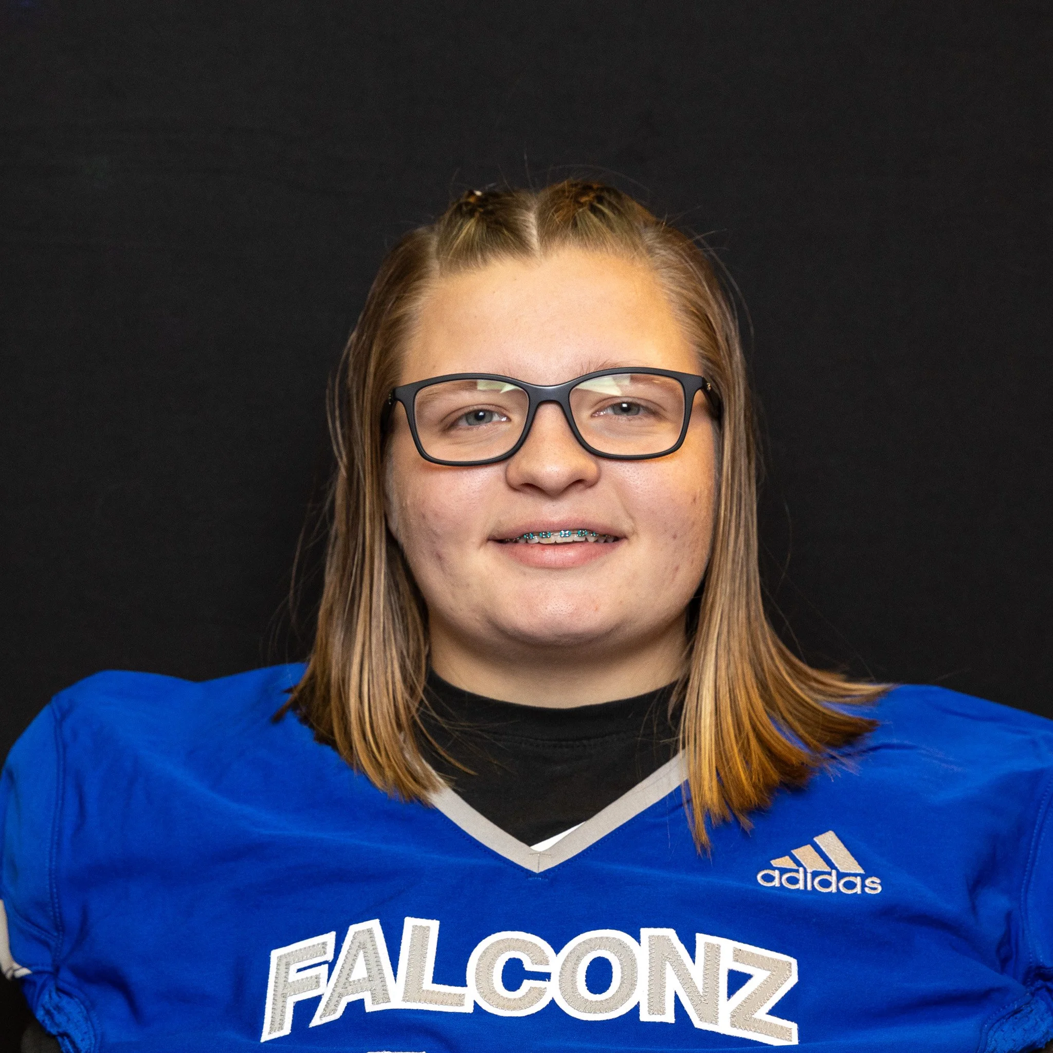 Young football player with shoulder-length hair wearing glasses, a blue football jersey with white text 'FALCONZ', and braces, smiling at the camera against a black background.