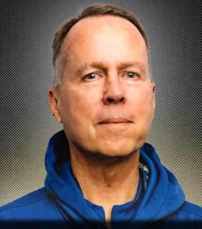 A portrait of a middle-aged man with short light brown hair, wearing a blue jacket, against a gradient dark background.