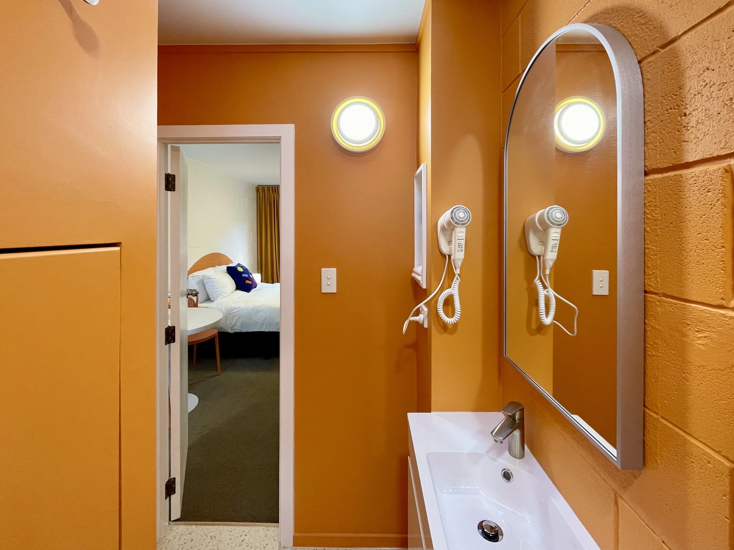 Bathroom with orange walls, a wall-mounted mirror, a white sink, wall-mounted hairdryer, and a bedroom visible through the doorway.