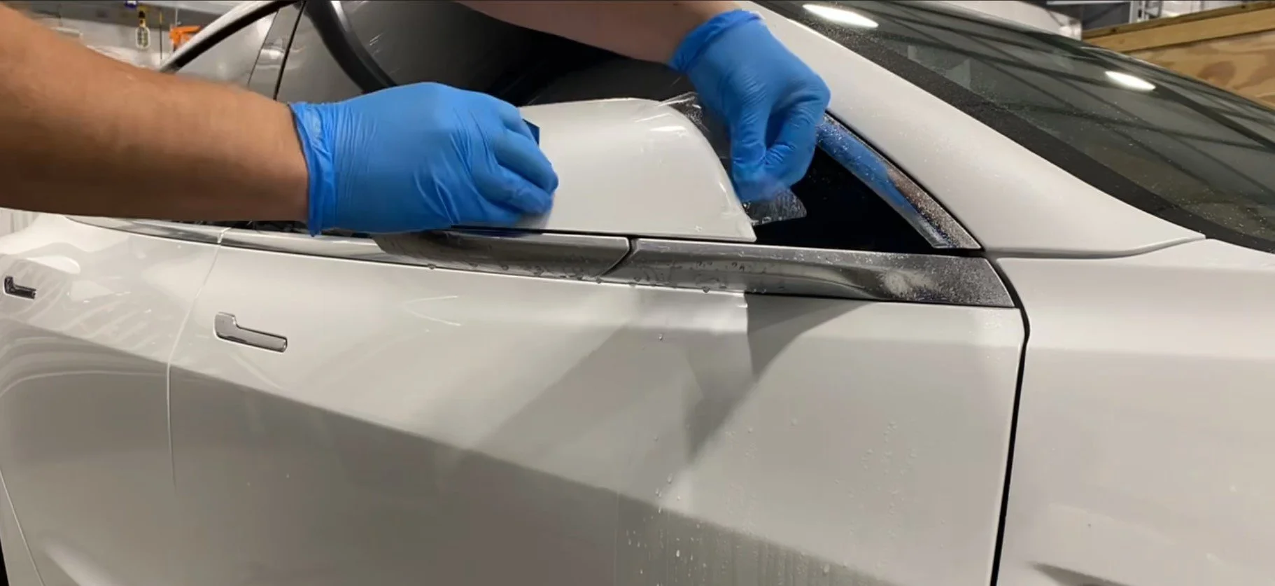 Person wearing blue gloves applying a protective film to the front of a white car.