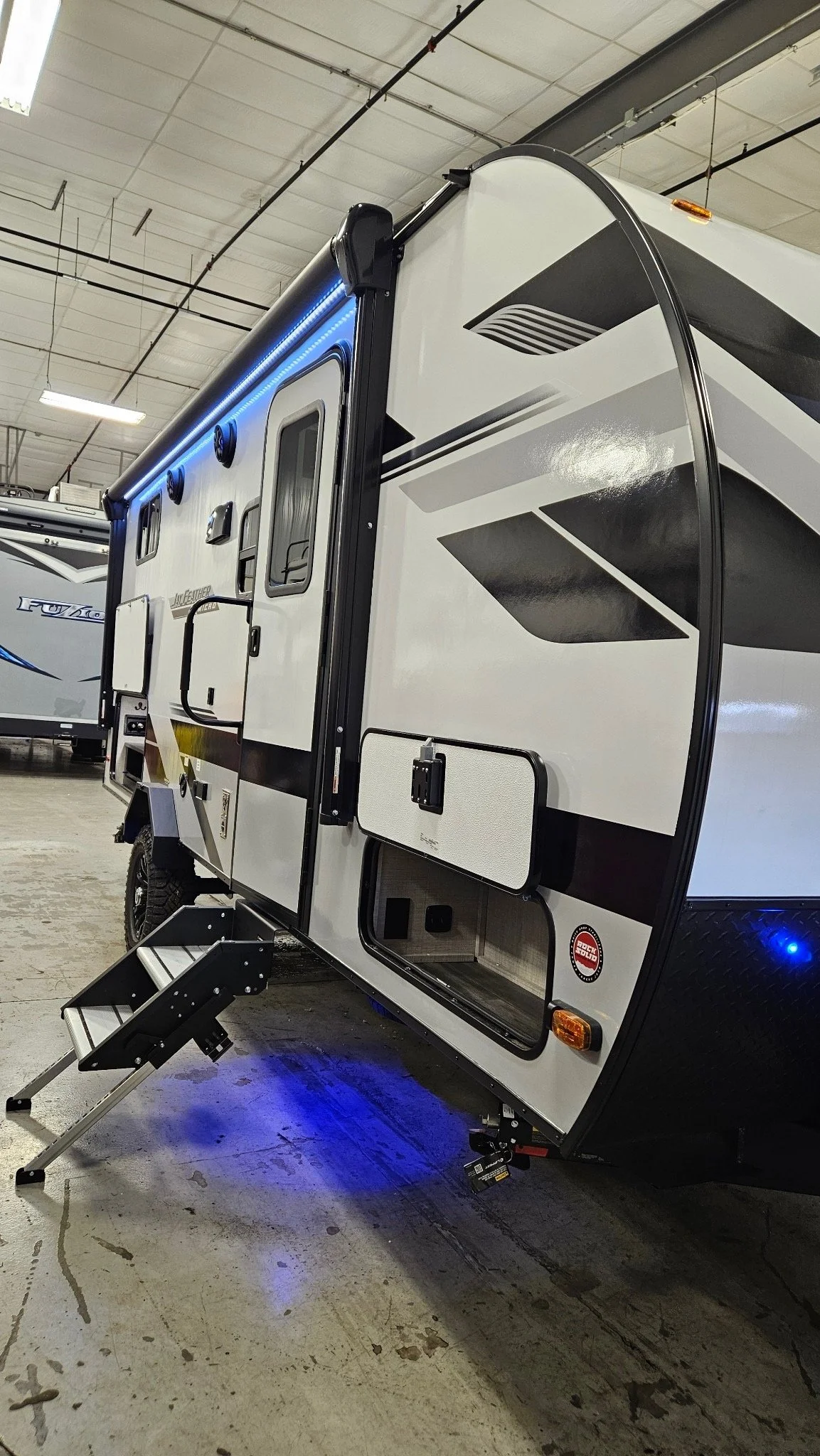 A modern travel trailer or RV with black and white exterior, blue LED lighting, and steps leading to the door, inside a warehouse or dealership.