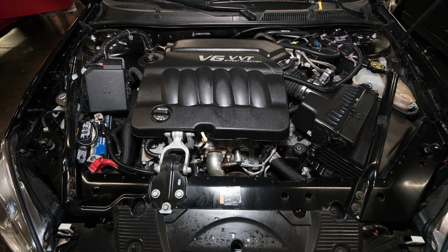 View of a car's engine bay with a V6 VVT engine cover, various hoses, wires, a battery, and fluid reservoirs.