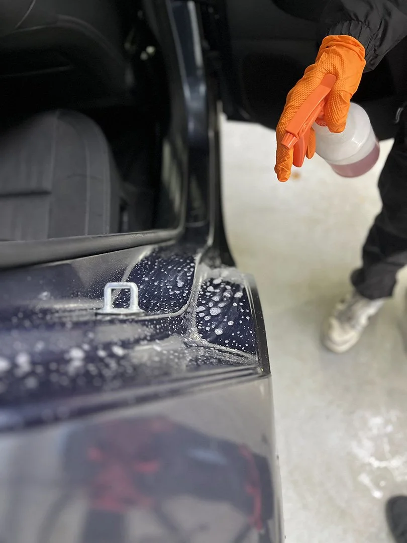 Person wearing orange gloves holding a spray bottle, cleaning or washing the exterior of a black vehicle in a garage or workshop.