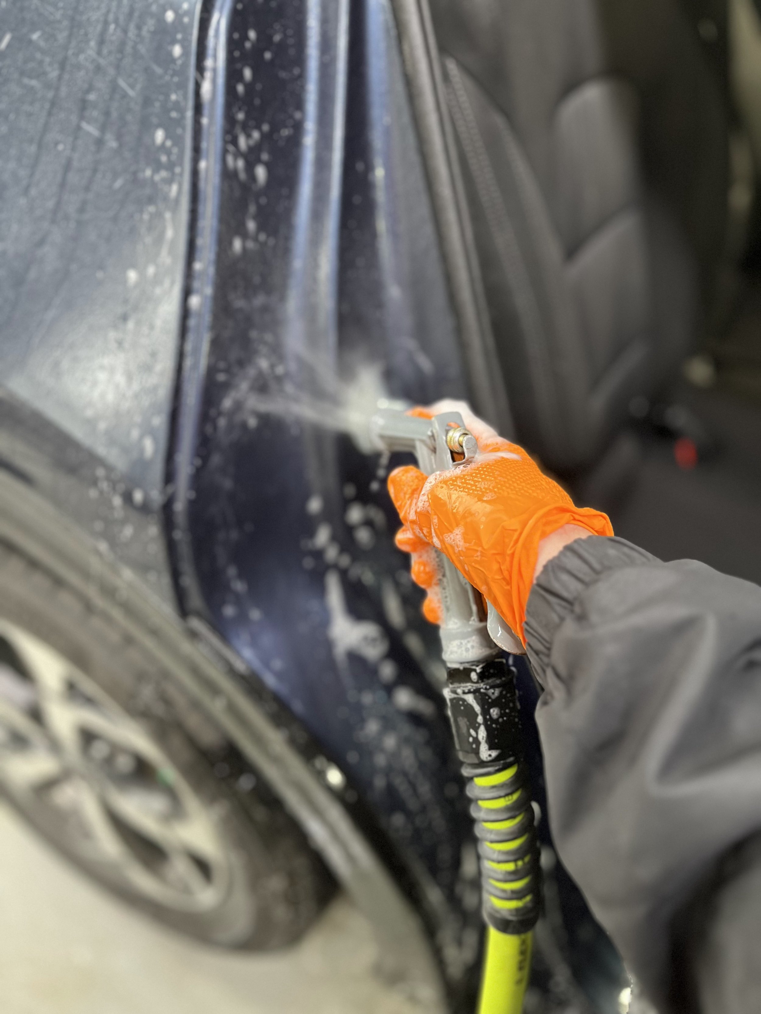 A person wearing orange gloves and a dark jacket is using a power washer to clean a black vehicle's exterior, with soap suds visible on the car's surface.