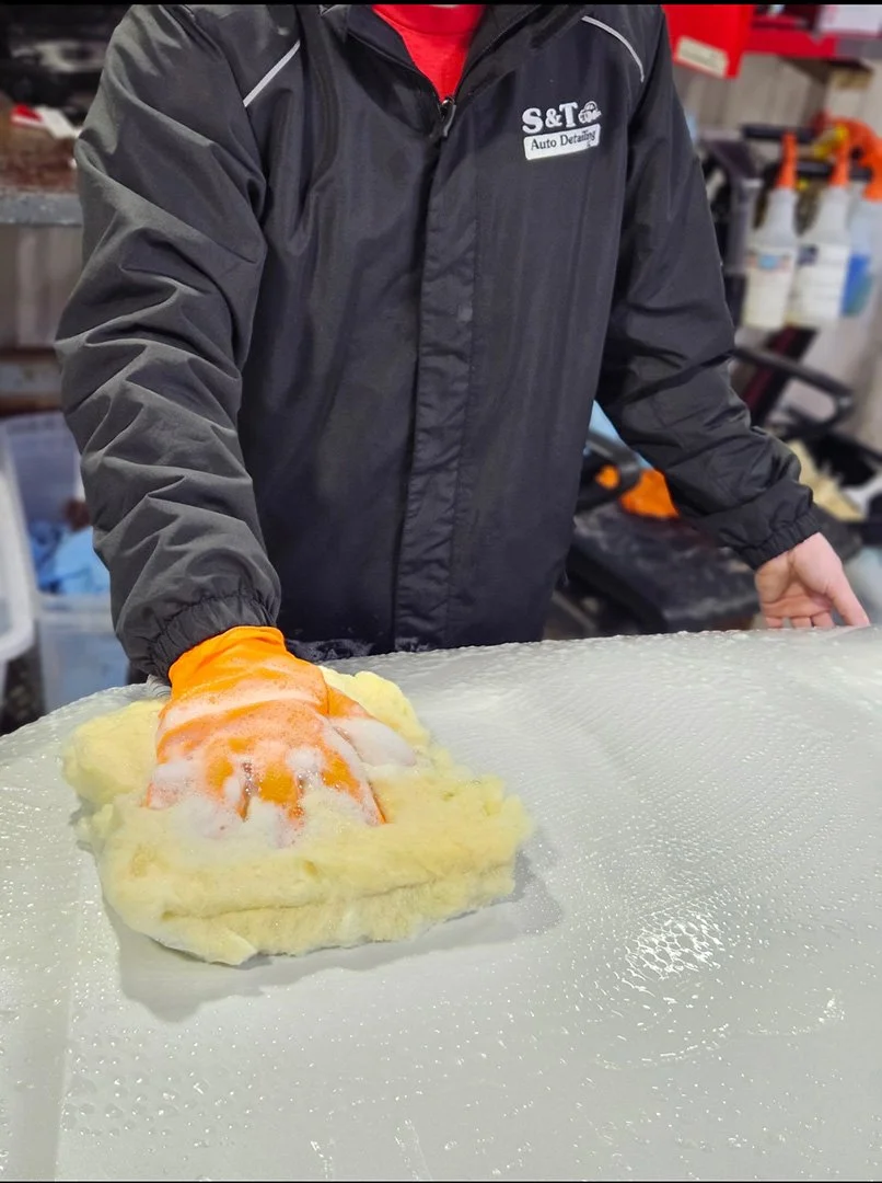 A person in an S&T Auto Detailing jacket wearing orange gloves is cleaning a large piece of sponge on a glass surface, with cleaning supplies visible in the background.