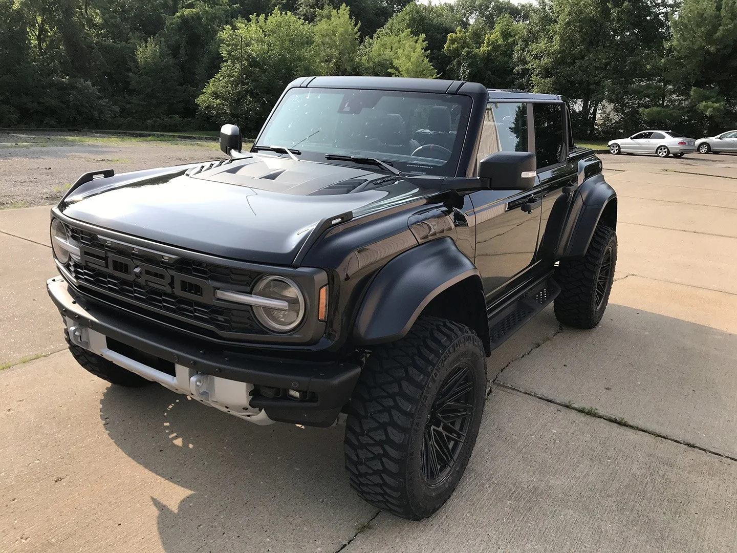 Black custom Ford Bronco parked on a concrete lot with trees in the background.