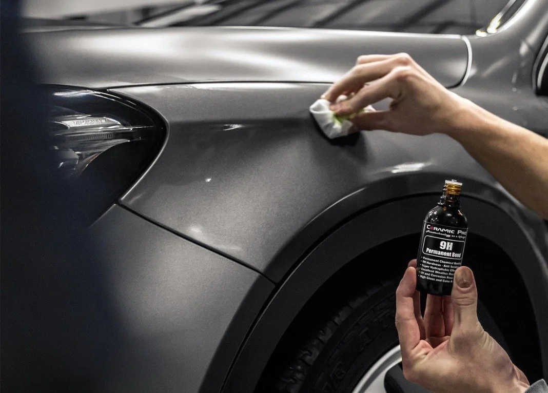 Person applying a chemical product on a car's matte black exterior with a cloth while holding a bottle labeled '9H Permanent Bond' for automotive paint protection.