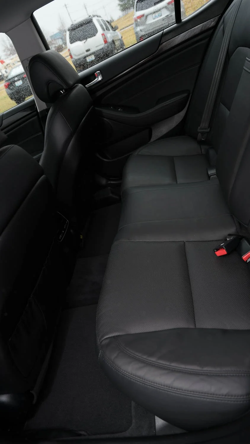 The image shows the backseat area of a car with black leather seats, a window with city view and rain, and multiple vehicles outside.