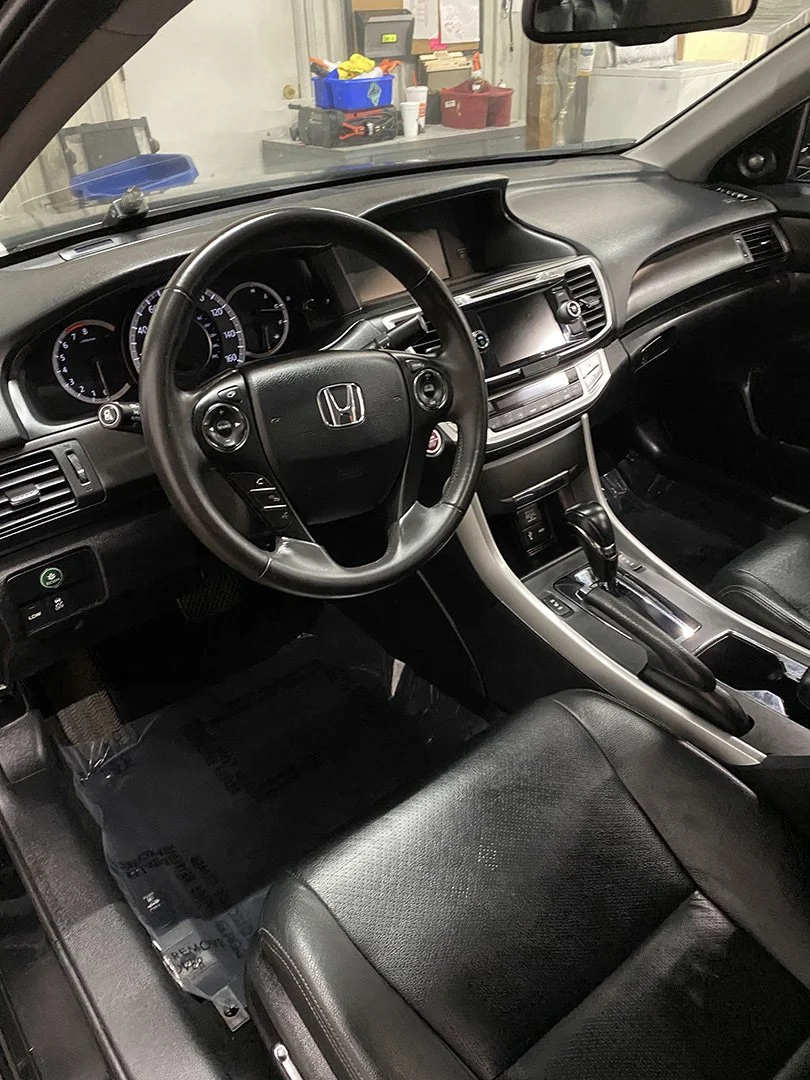 Interior of a Honda car, showing the steering wheel, dashboard, gear shift, and front seats, with a view of a garage or workshop in the background.