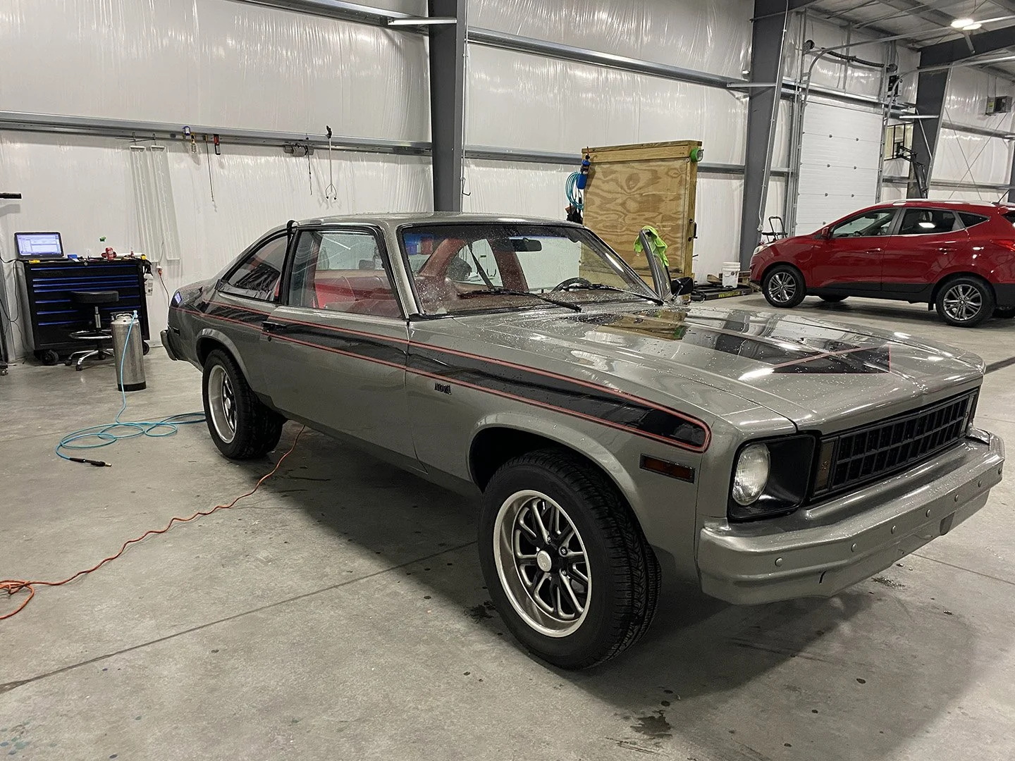 A vintage gray and black sports car with red pinstripes is parked inside a garage, with a modern red car in the background. The garage features a mechanic's work area with tools, a toolbox, and an air compressor connected by hoses.