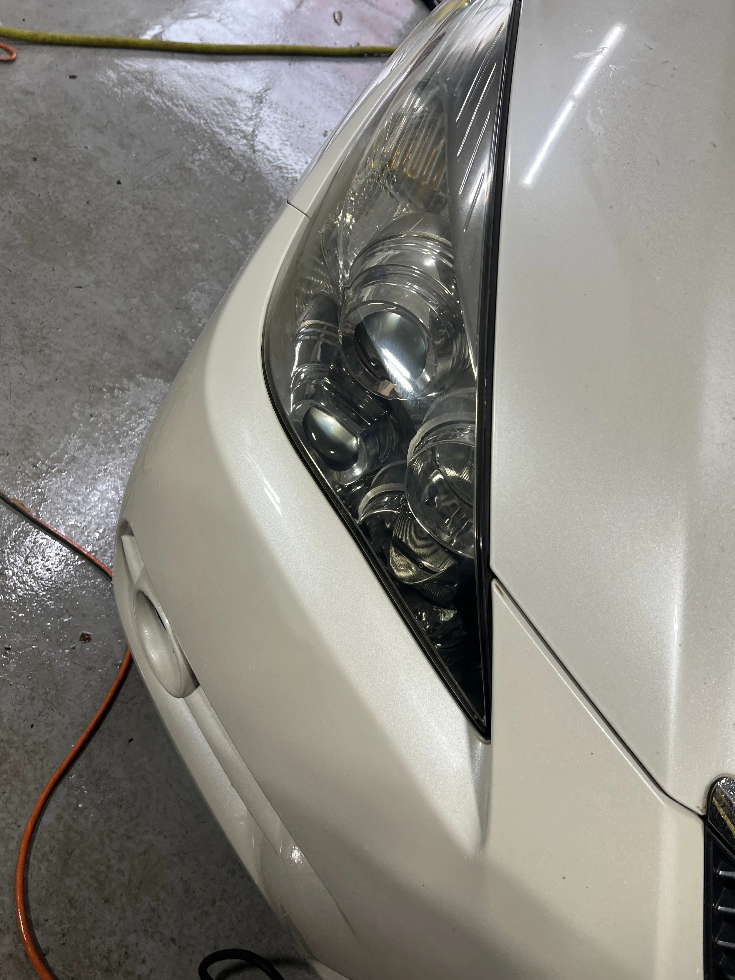 Close-up of the front right headlight of a white car, showing the clear lens and internal projector elements, with a wet concrete floor and an orange extension cord in the background.