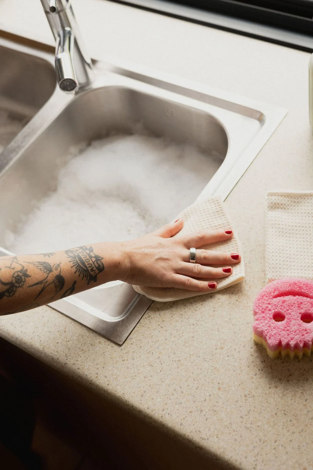 A cleaner from Another Mother wipes a kitchen bench beside a sink full of soapy water, demonstrating the practical, non-judgemental housekeeping services offered to Perth families.