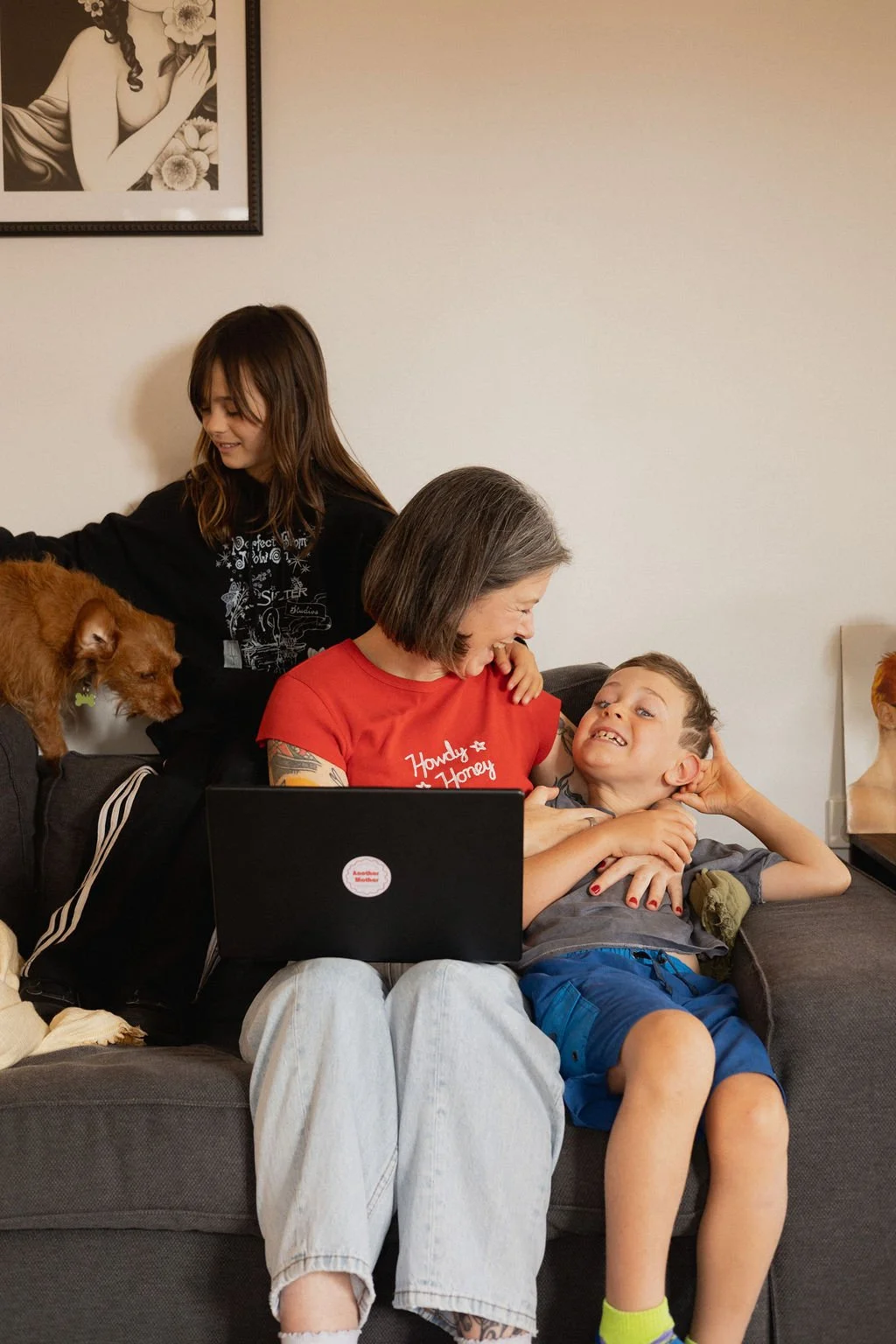 Hannah Purvey-Tyrer, founder of Another Mother, sits on the couch in a Perth home working on her laptop with her children and family dog beside her, reflecting the warm, family-focused support behind her non-judgemental housekeeping service.