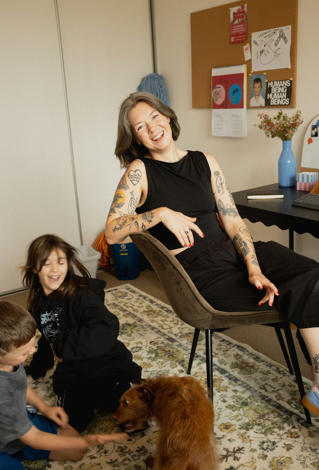 The founder of Another Mother sits smiling in a warm, lived-in home office while children play beside her, capturing the supportive, family-centred ethos behind the Perth-based non-judgemental housekeeping service.