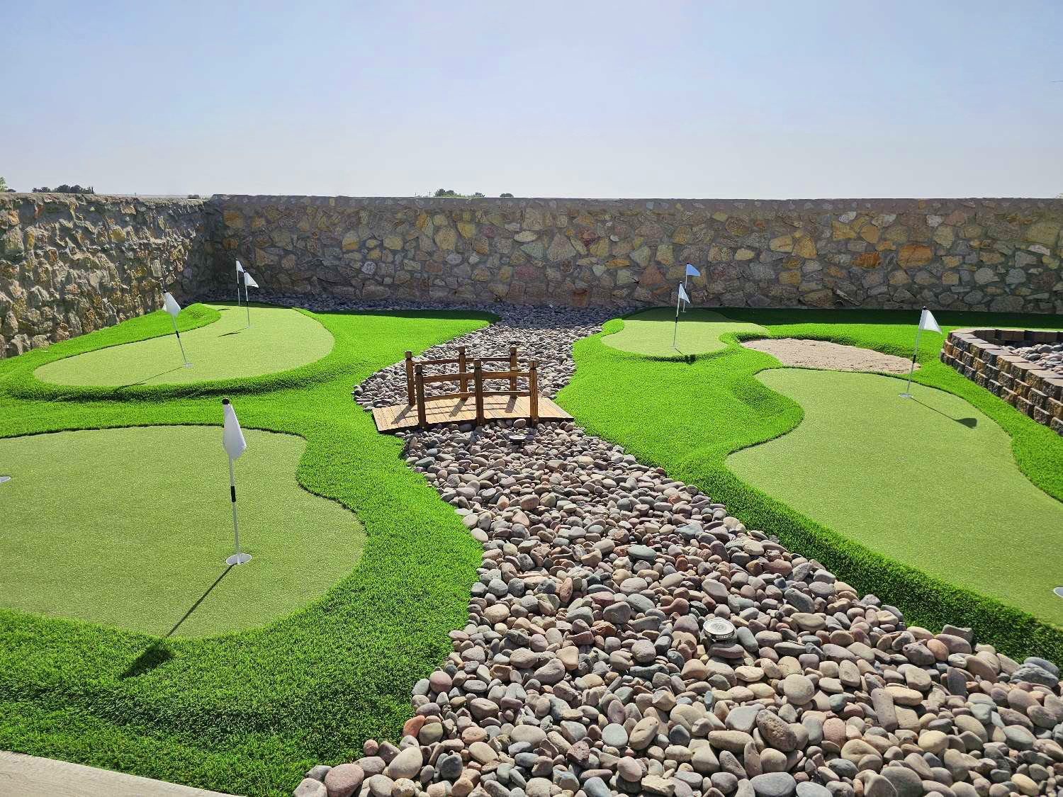 Miniature golf course with four holes, small bridge, pebble pathway, and stone wall background under clear sky.
