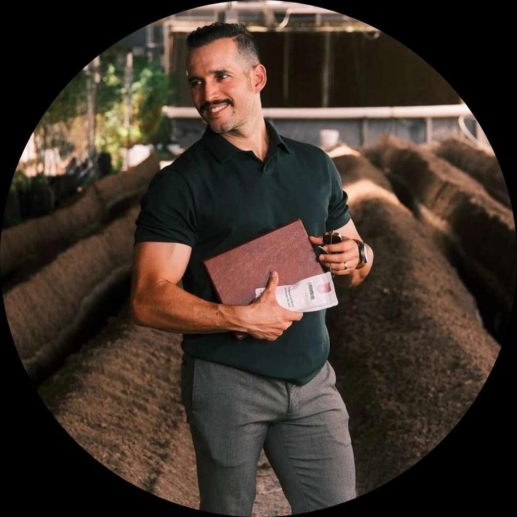 A man with short dark hair and a beard, wearing a black polo shirt and gray pants, standing in a greenhouse or plant nursery, holding a brown folder and a glass of wine, smiling and looking to the side.