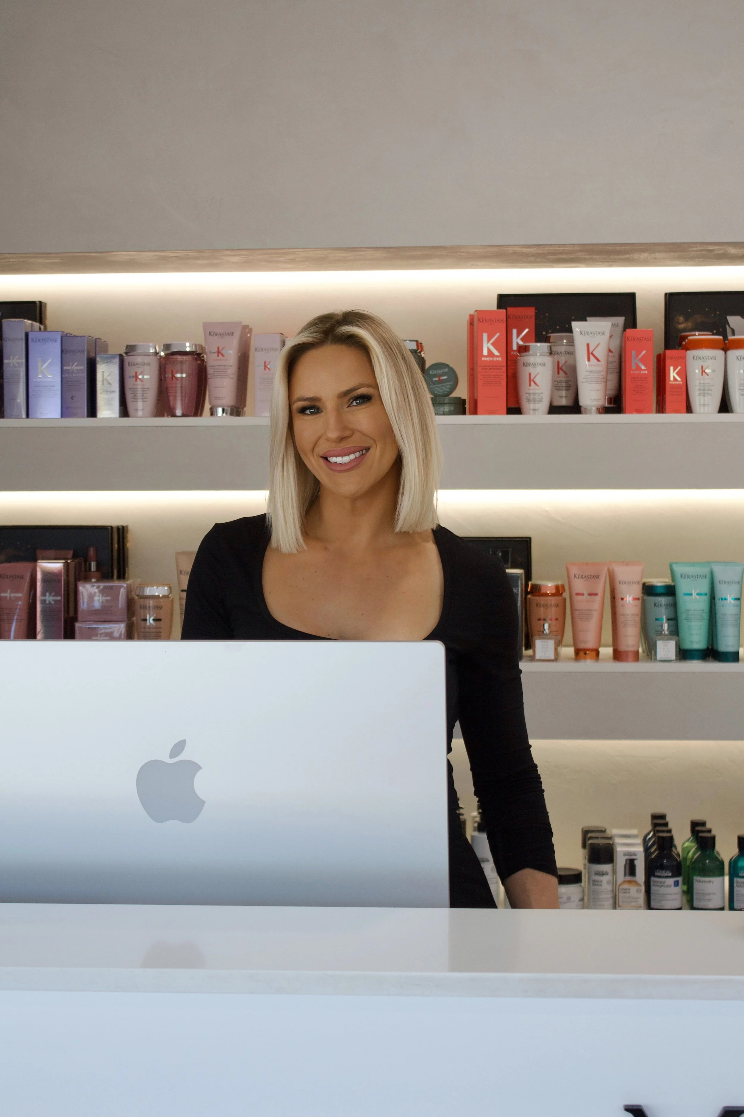 Smiling woman with blonde hair sitting behind a white desk in front of an Apple iMac, with shelves of skincare products in the background.