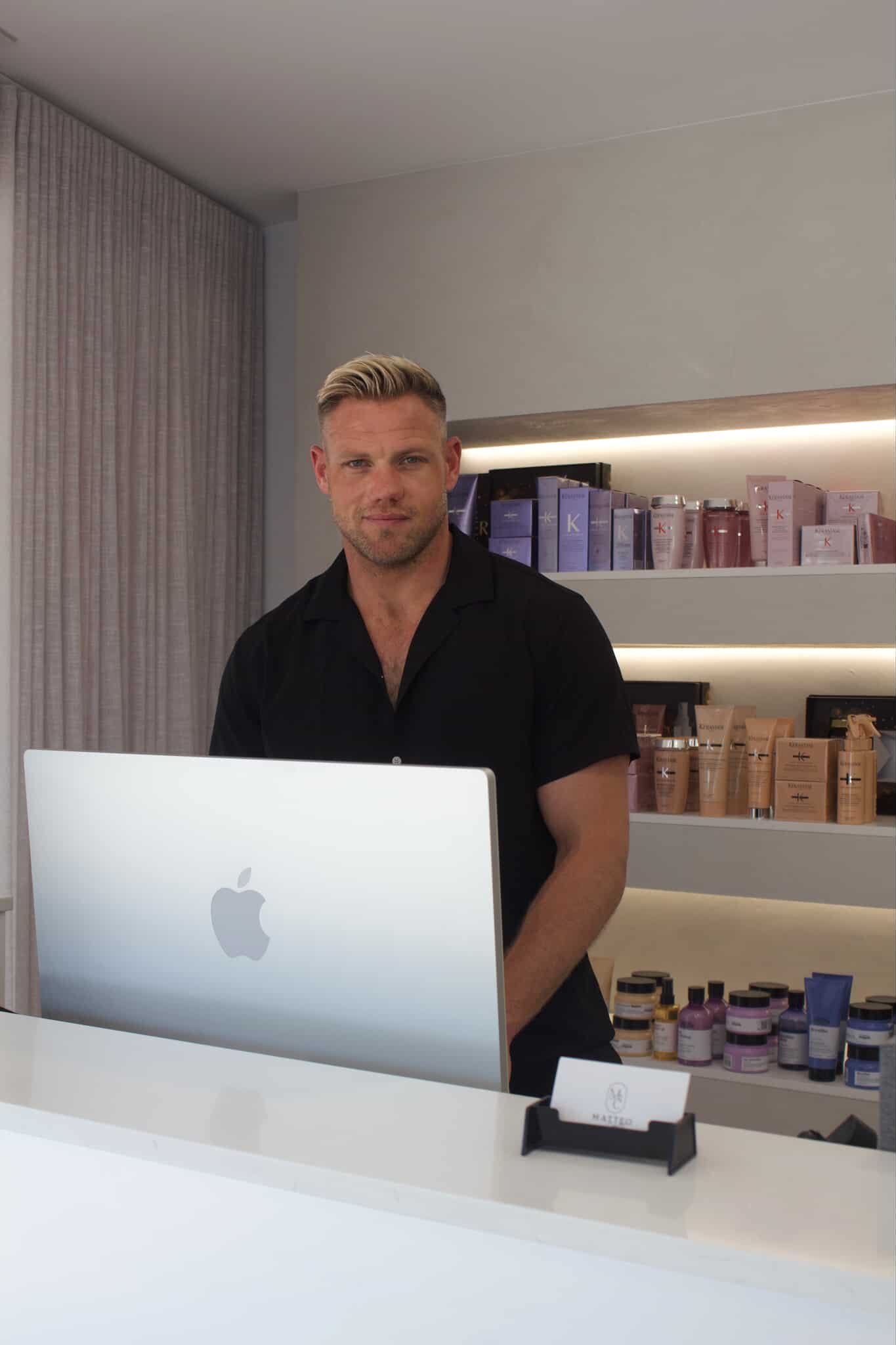 A man with blonde hair and a beard standing behind a white reception desk with an Apple iMac. Shelves with skincare products are in the background.