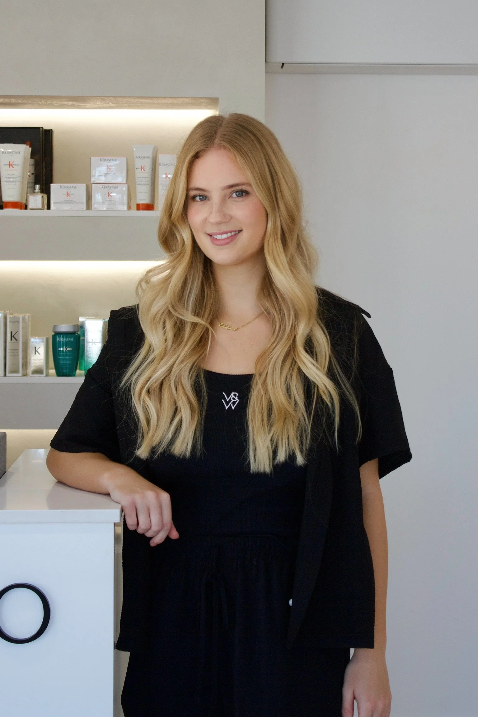 A young woman with long blonde wavy hair, wearing a black outfit, stands in front of a shelf of hair care products.