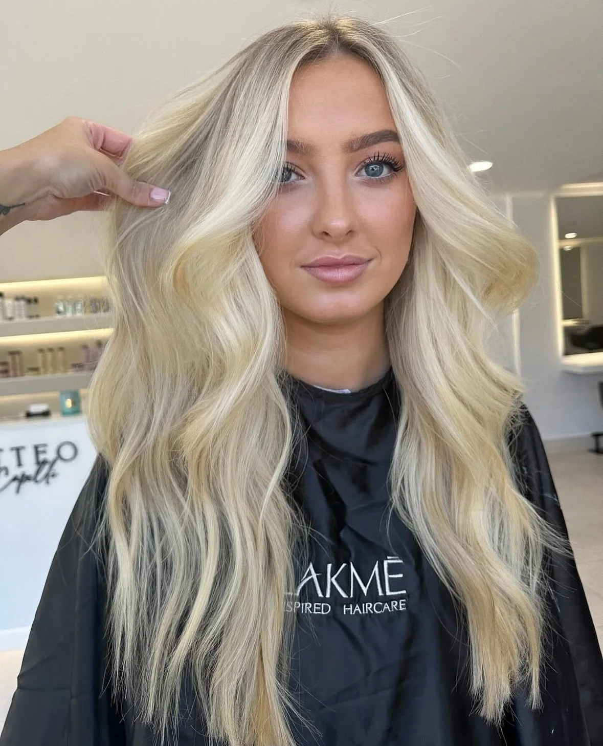 Young woman with long, wavy blonde hair sitting in a salon, wearing a black cape. Someone is holding a section of her hair, possibly getting a haircut or styling. The salon background includes shelves and mirrors.