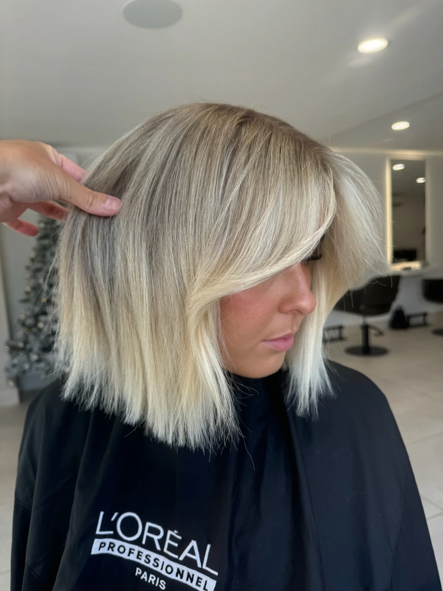 A woman with shoulder-length blonde hair sitting in a salon chair, wearing a black L'Oreal Professionnel cape inside a hair salon.