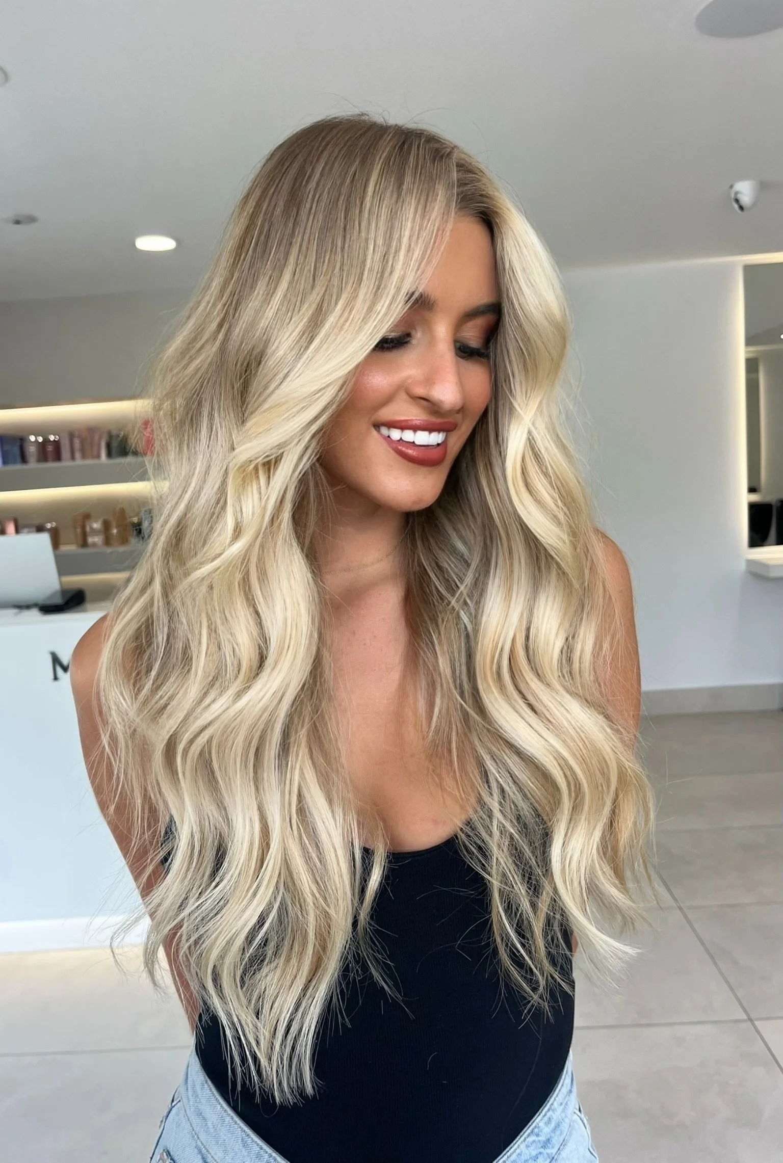 A woman with long, wavy blonde hair smiling in a modern indoor setting.