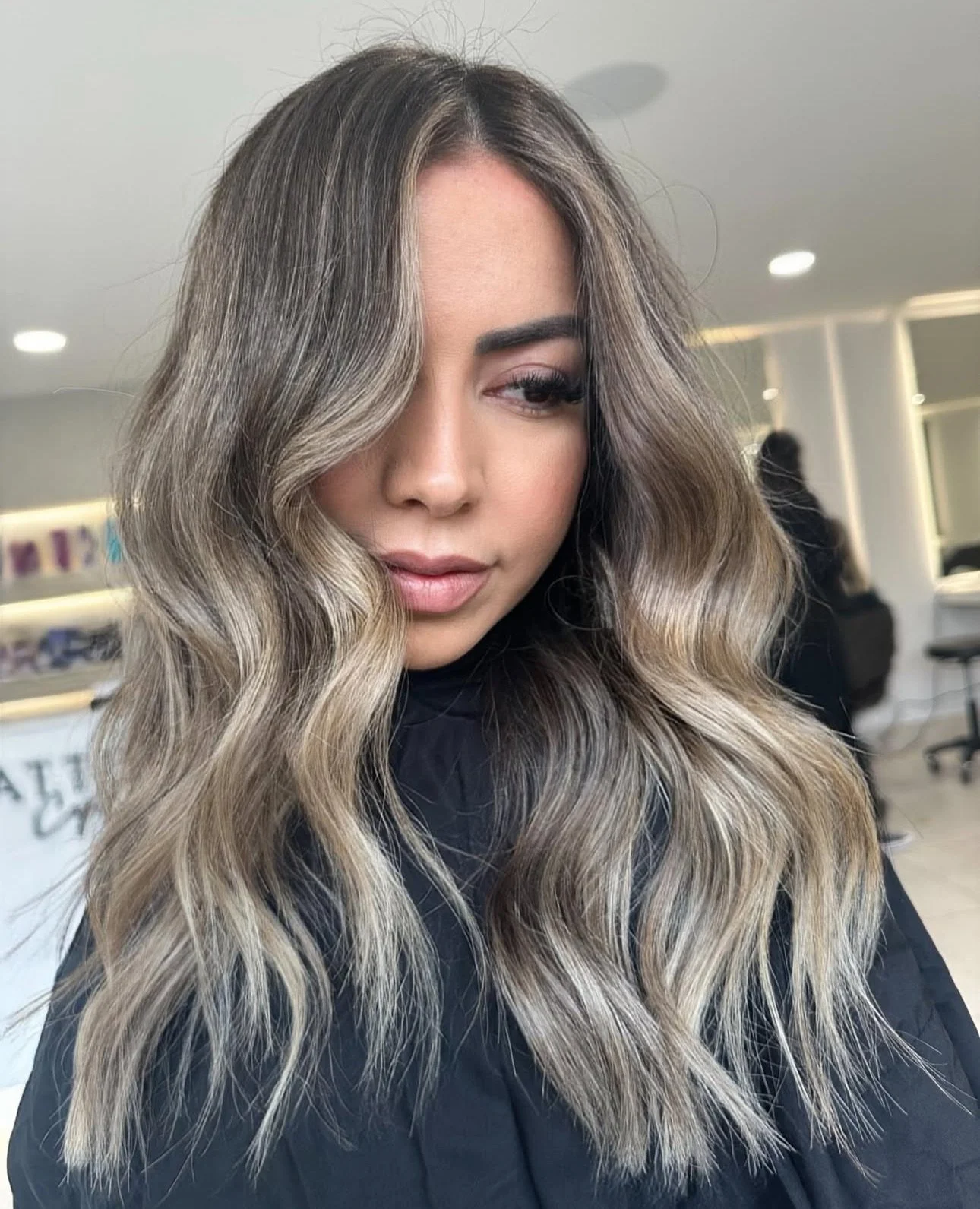Woman with long wavy hair in shades of blonde, brown, and ash, sitting in a salon.