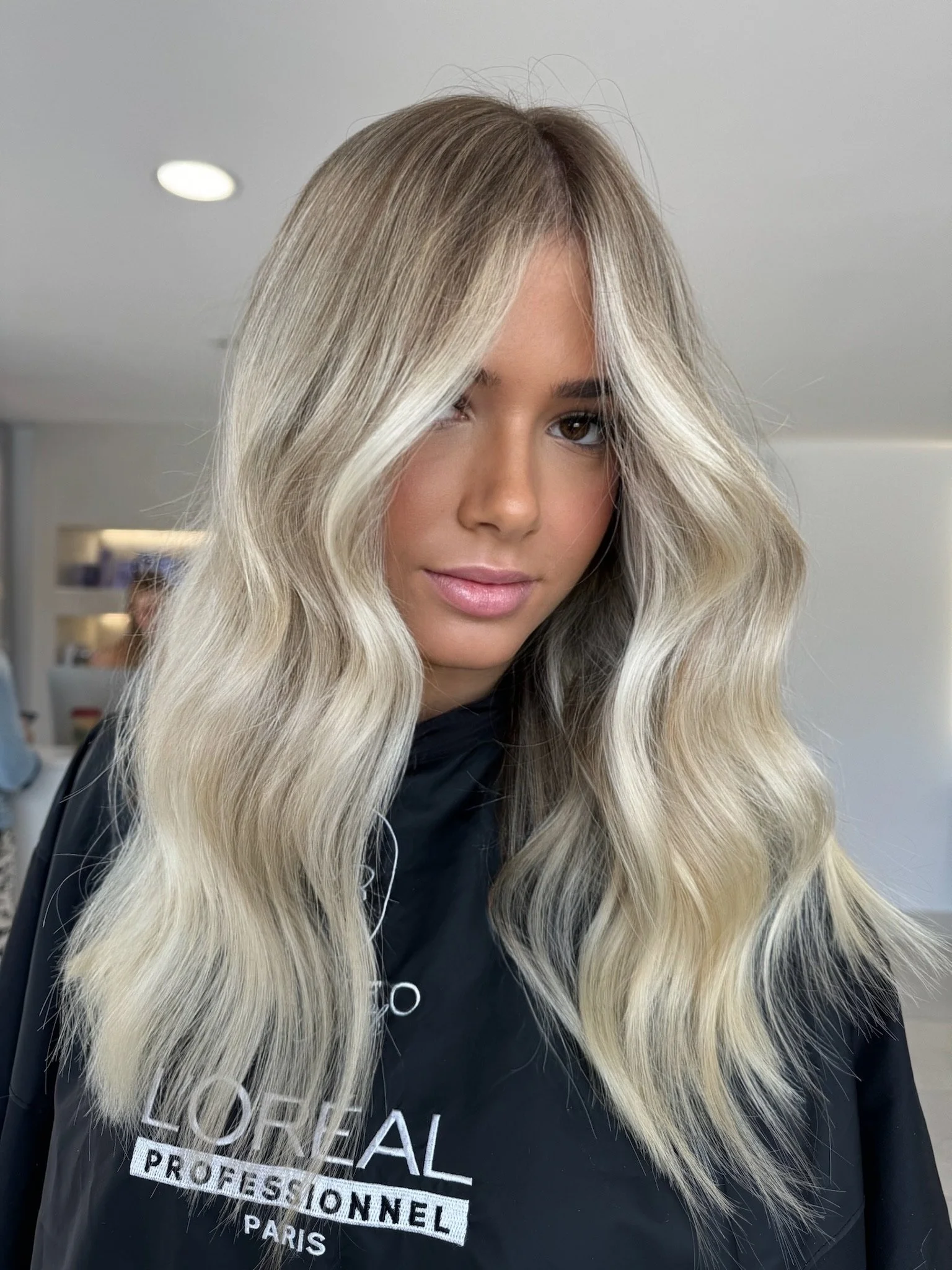 A woman with long, wavy ash blonde hair with darker roots, wearing a black cape with 'L'Oreal Professionnel Paris' logo, standing in a salon.