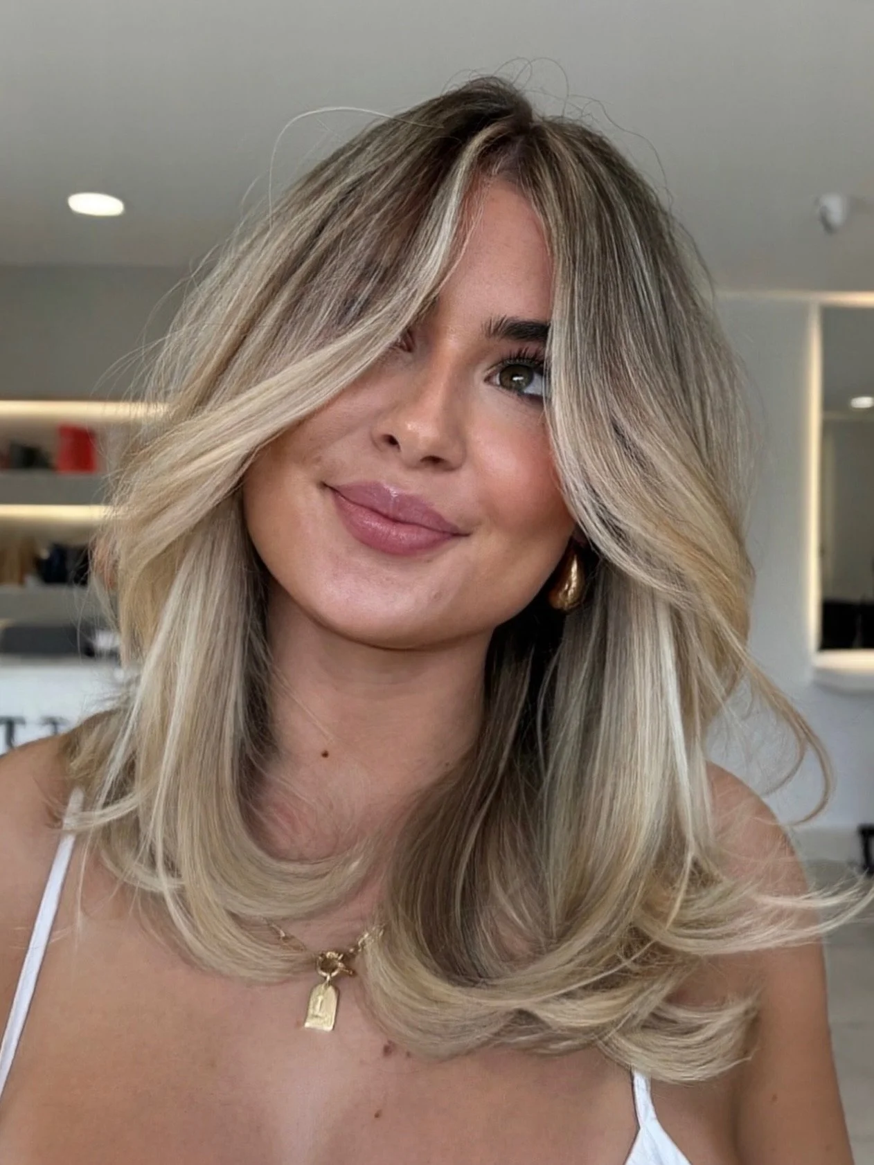 A young woman with long, wavy blonde hair and a slight smile, wearing a white top and gold jewelry, posing in an indoor setting.